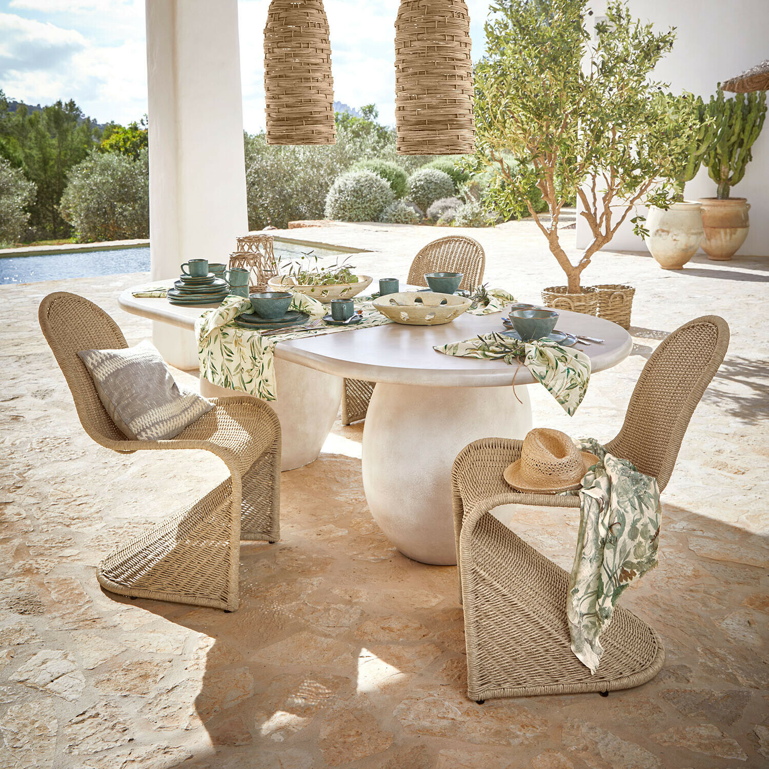 Stůl Arevalon Oválný stůl s bílou deskou a přírodními proutěnými židlemi na terase s výhledem do zahrady