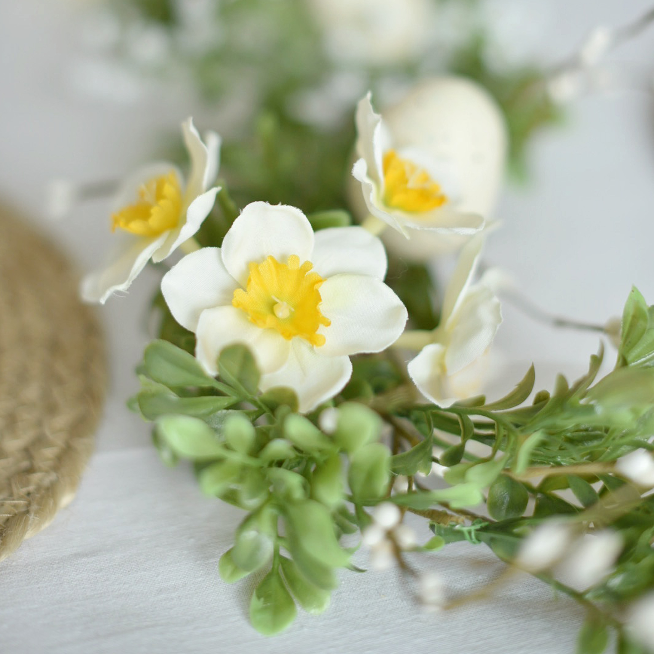 Dekorativní věnec s bílými a žlutými květy a zelenými listy na světlém pozadí