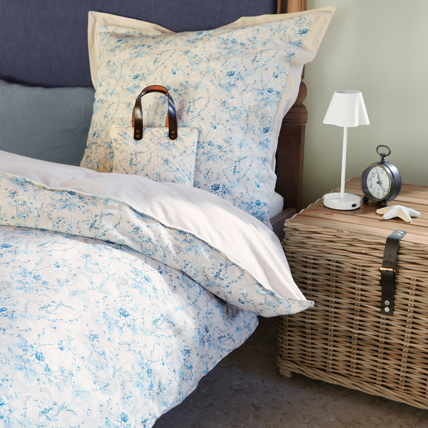 Bavlněné povlečení s jemným modrým květinovým vzorem na posteli vedle proutěné komody a lampy