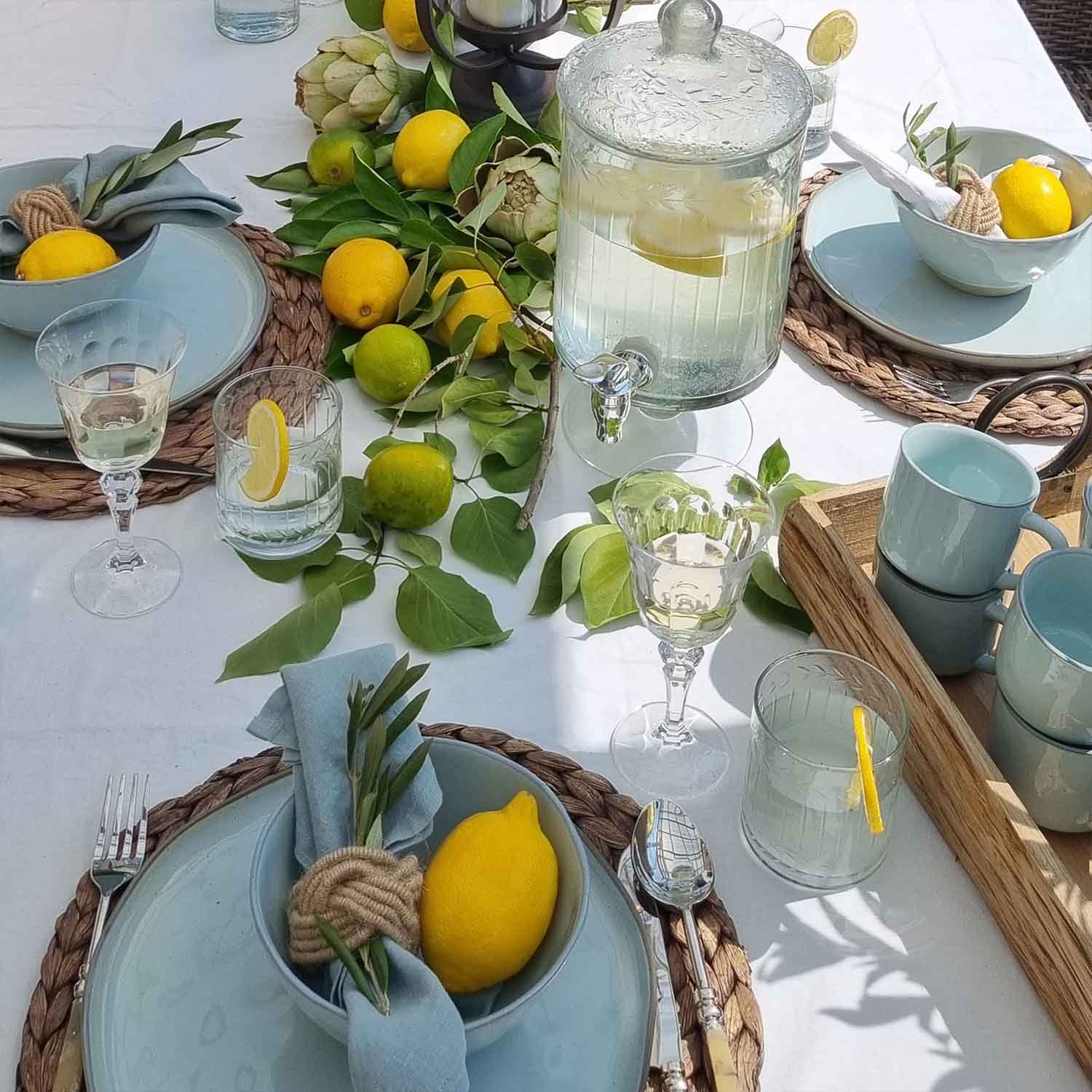 Letní stůl s modrým keramickým nádobím, citrony a zelenými listy na bílém ubrusu, dekorace stolu