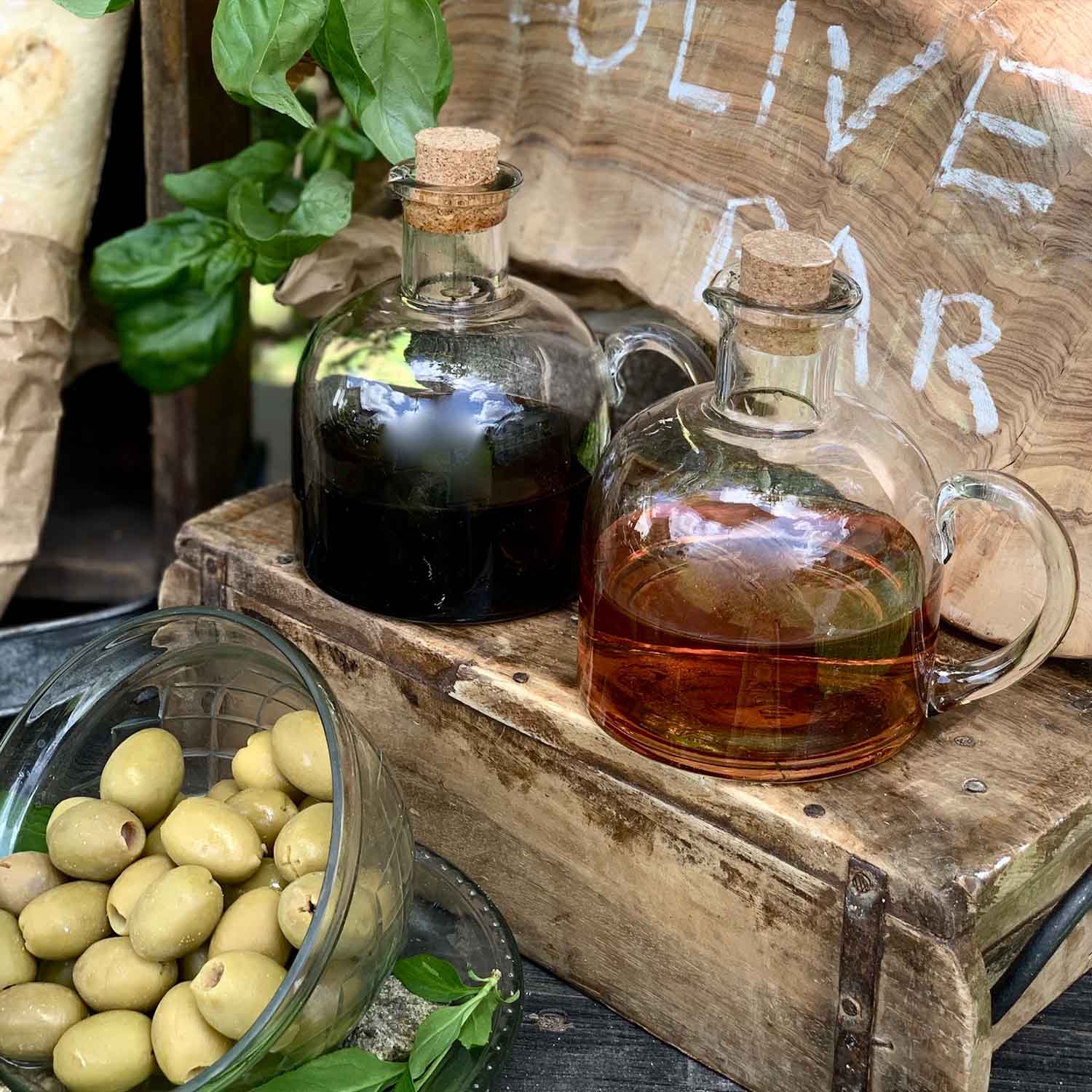 Skleněné lahve s korkem s olejem a octem na dřevěné bedně vedle zelených oliv a bazalkových listů