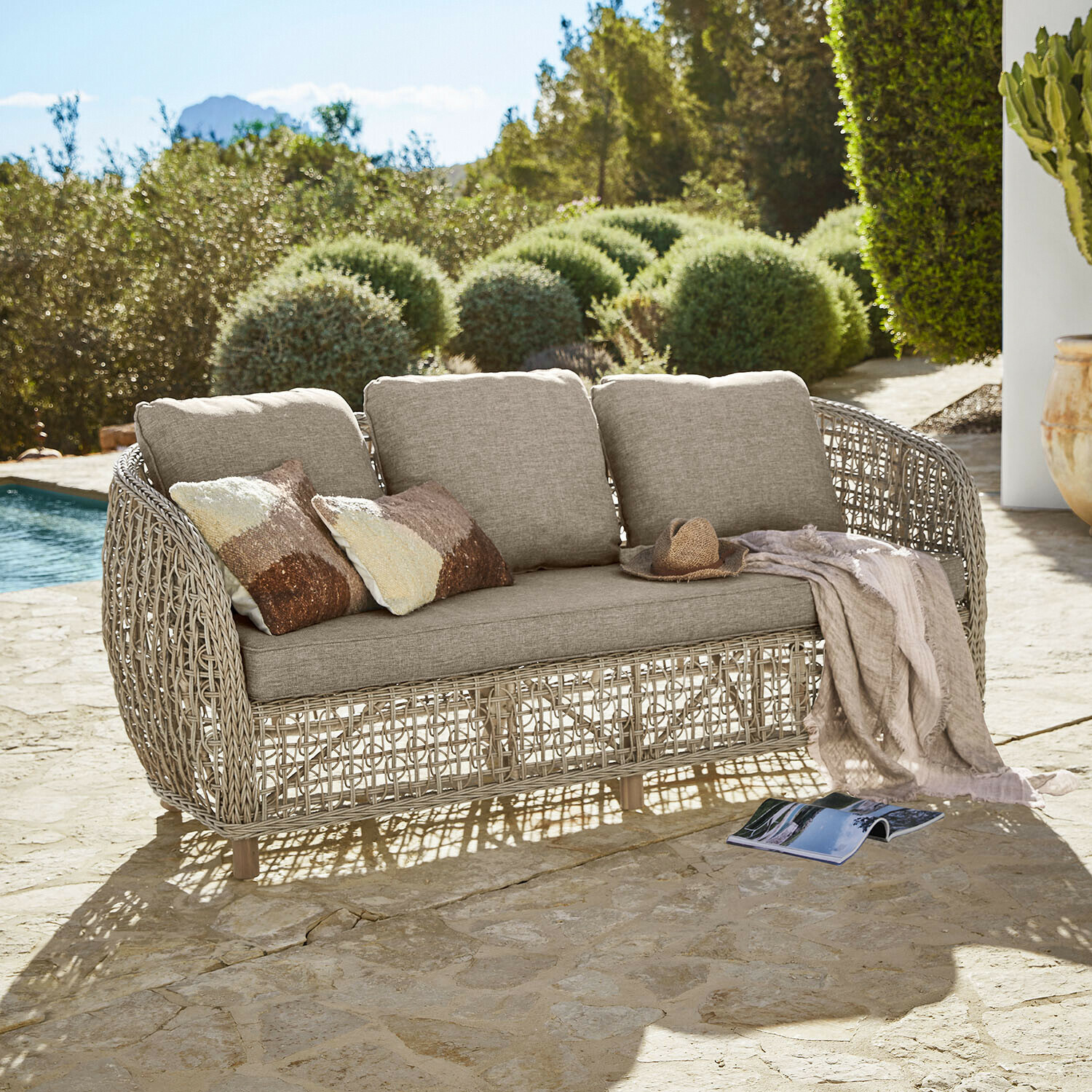 Zahradní pohovka s béžovými polštáři a ratanovým rámem na kamenné terase u bazénu