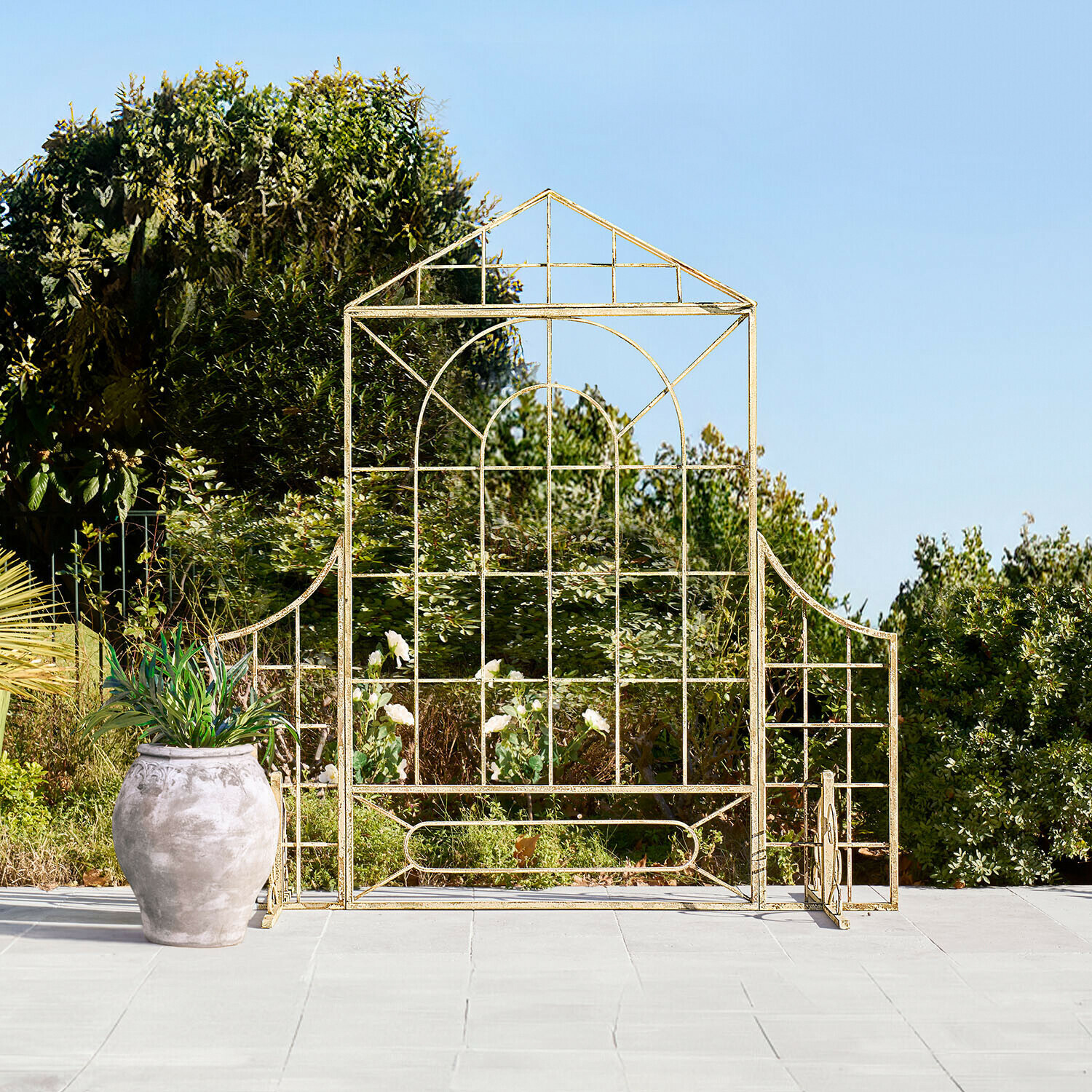 Zlatá kovová zahradní mříž ve tvaru domu na terase s květináčem a zelení v pozadí