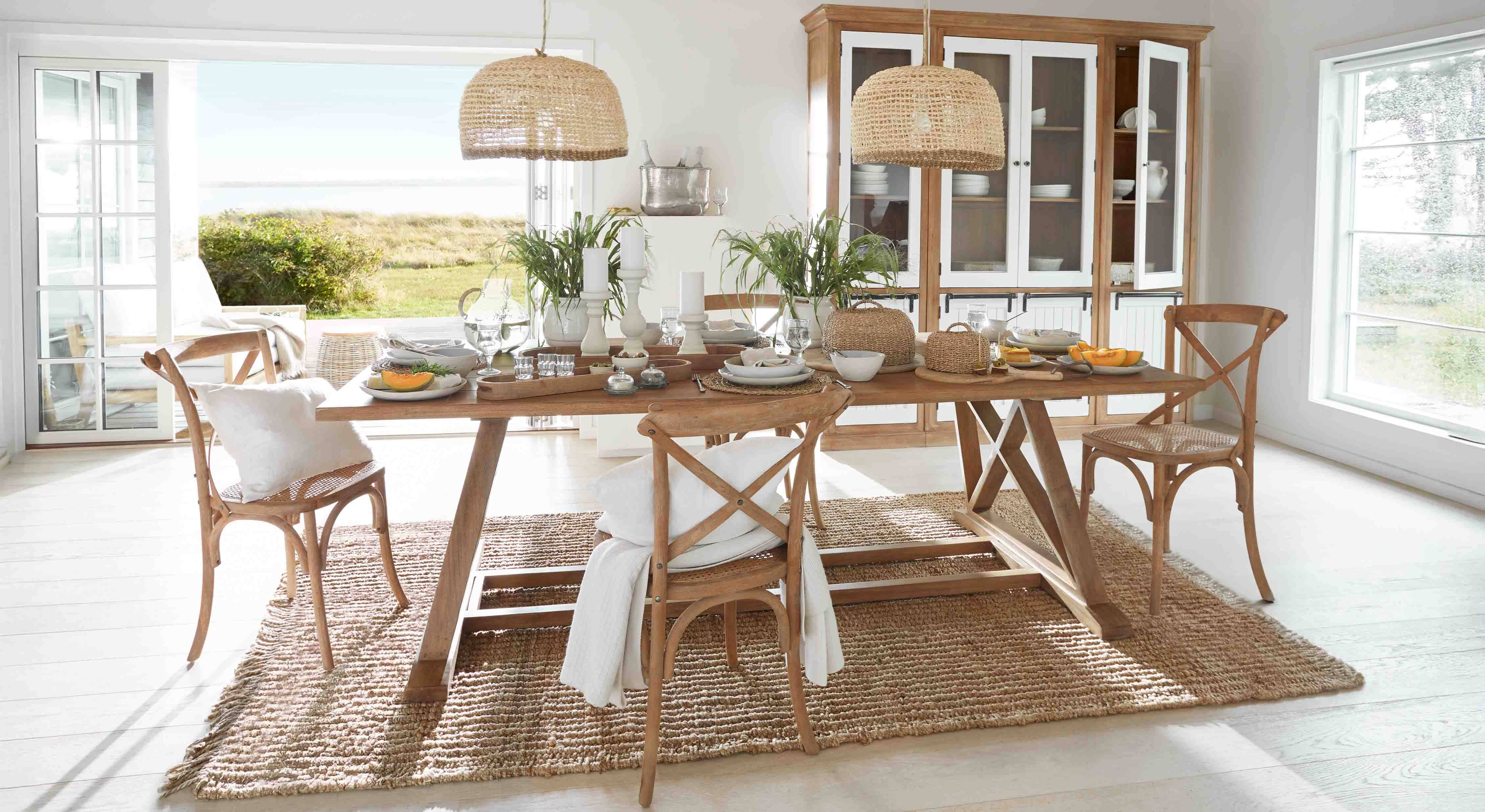 Jídelna s dřevěným stolem a ratanovými židlemi na přírodním koberci, rostlinná dekorace a proutěná svítidla