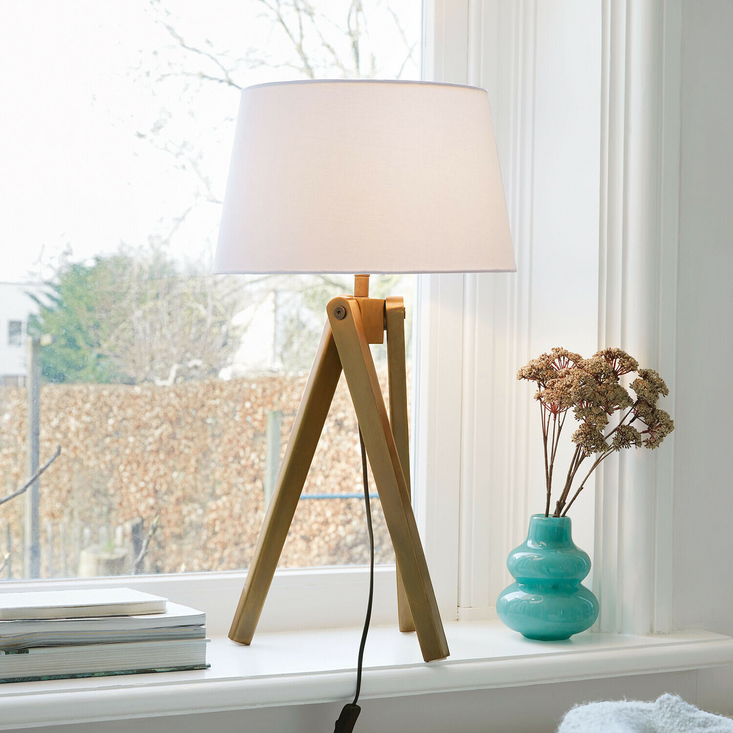 Stolní lampa Kelvio Stolní lampa s dřevěnou základnou a bílým stínidlem na parapetu vedle tyrkysové vázy