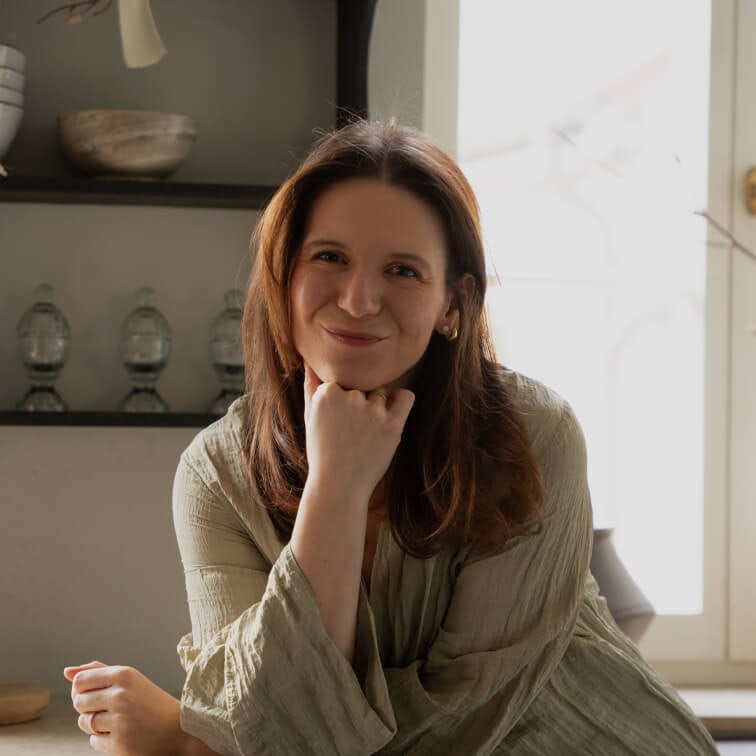 Usměvavá žena v zelené lněné košili sedí u stolu v světlém interiéru