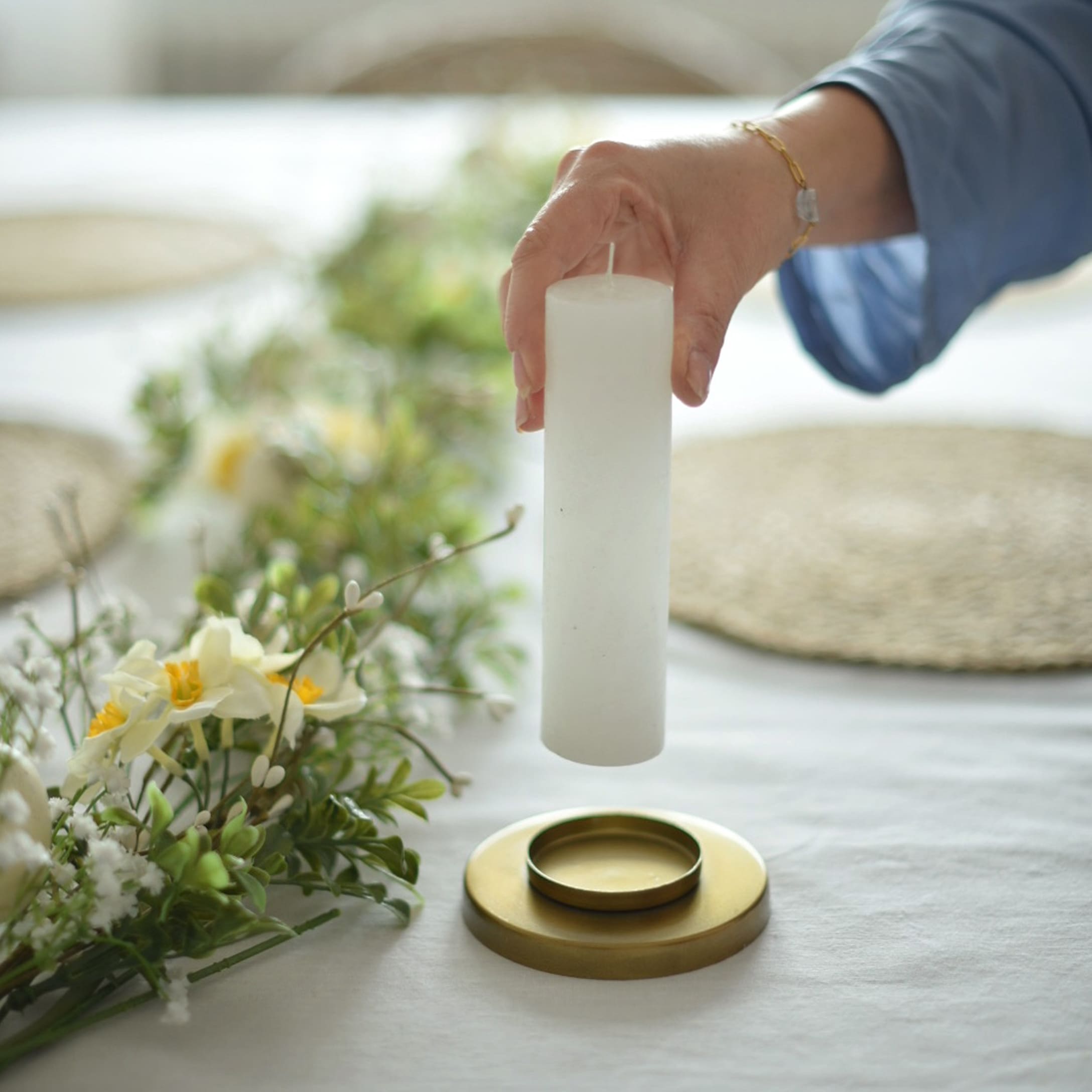 Bílá válecová svíčka držená nad zlatým svícnem s květinovou dekorací na bílém ubrusu