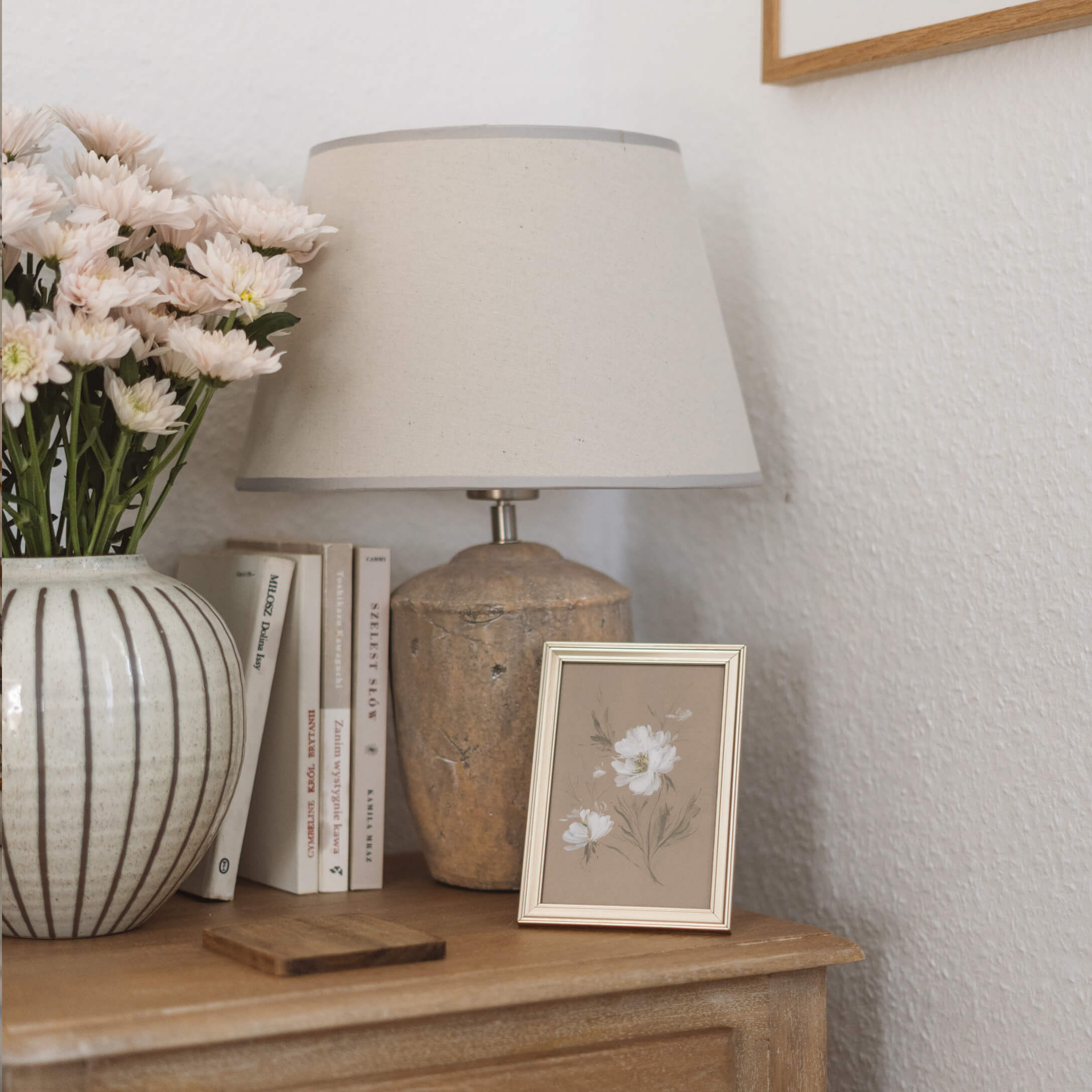 Stolní lampa s béžovým stínidlem a keramickou základnou na dřevěném stolku s květinami a knihami