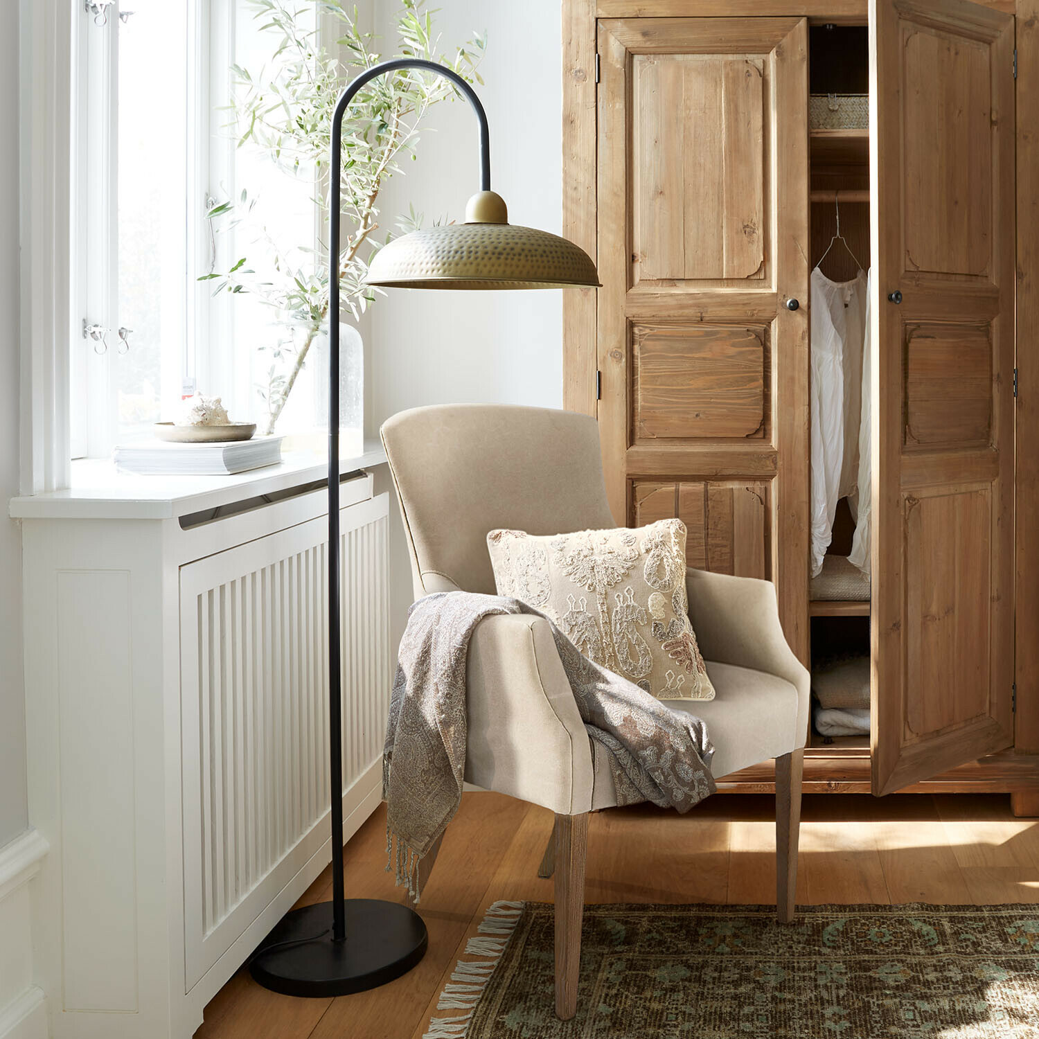 Béžové křeslo s dřevěnými nohami, ozdobný polštář a deka, vedle stojací kovová lampa s mosazným stínidlem