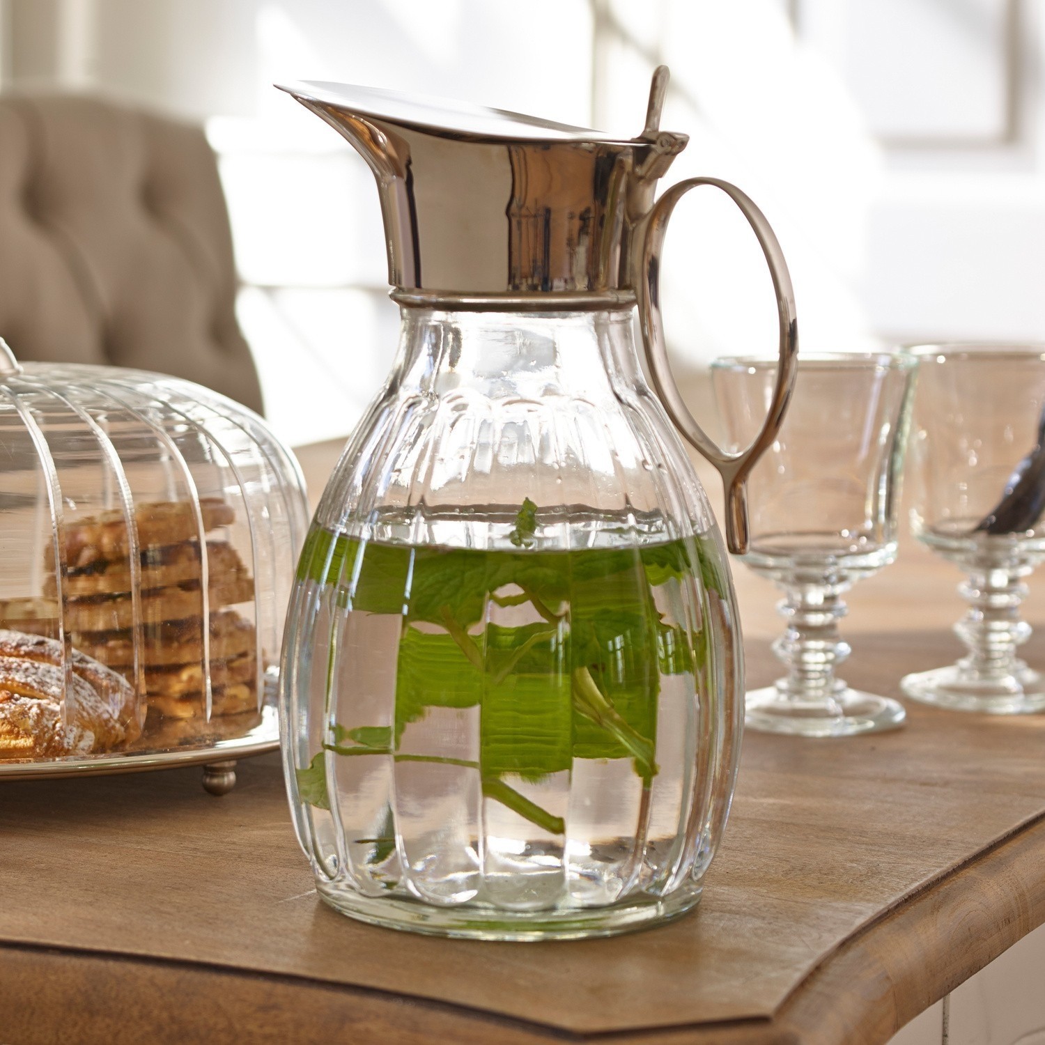 Džbán Agudelle Skleněná karafa s kovovým víkem a rukojetí, voda s mátovými listy na stole s pečivem