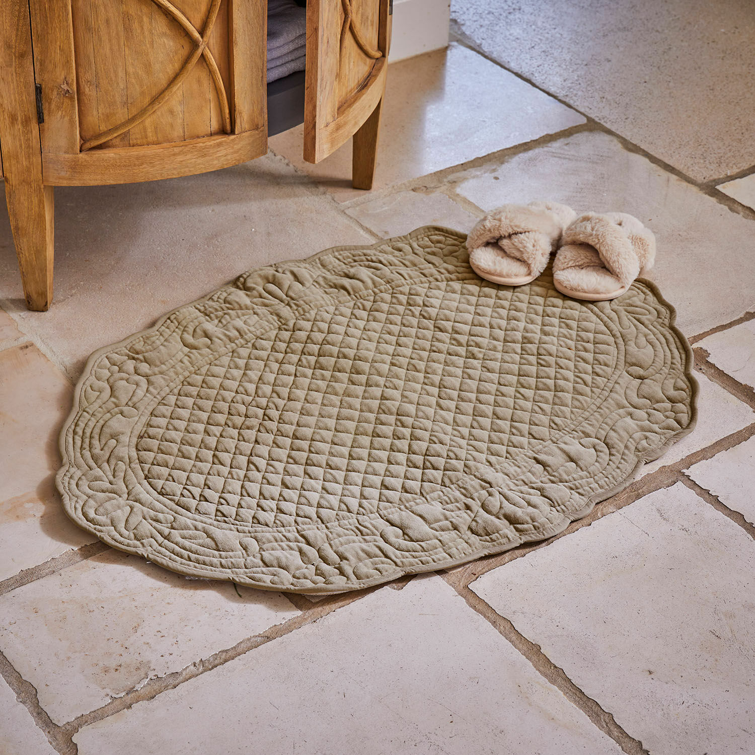 Oválný prošívaný bavlněný koberec olivové barvy na kamenné podlaze s béžovými pantoflemi