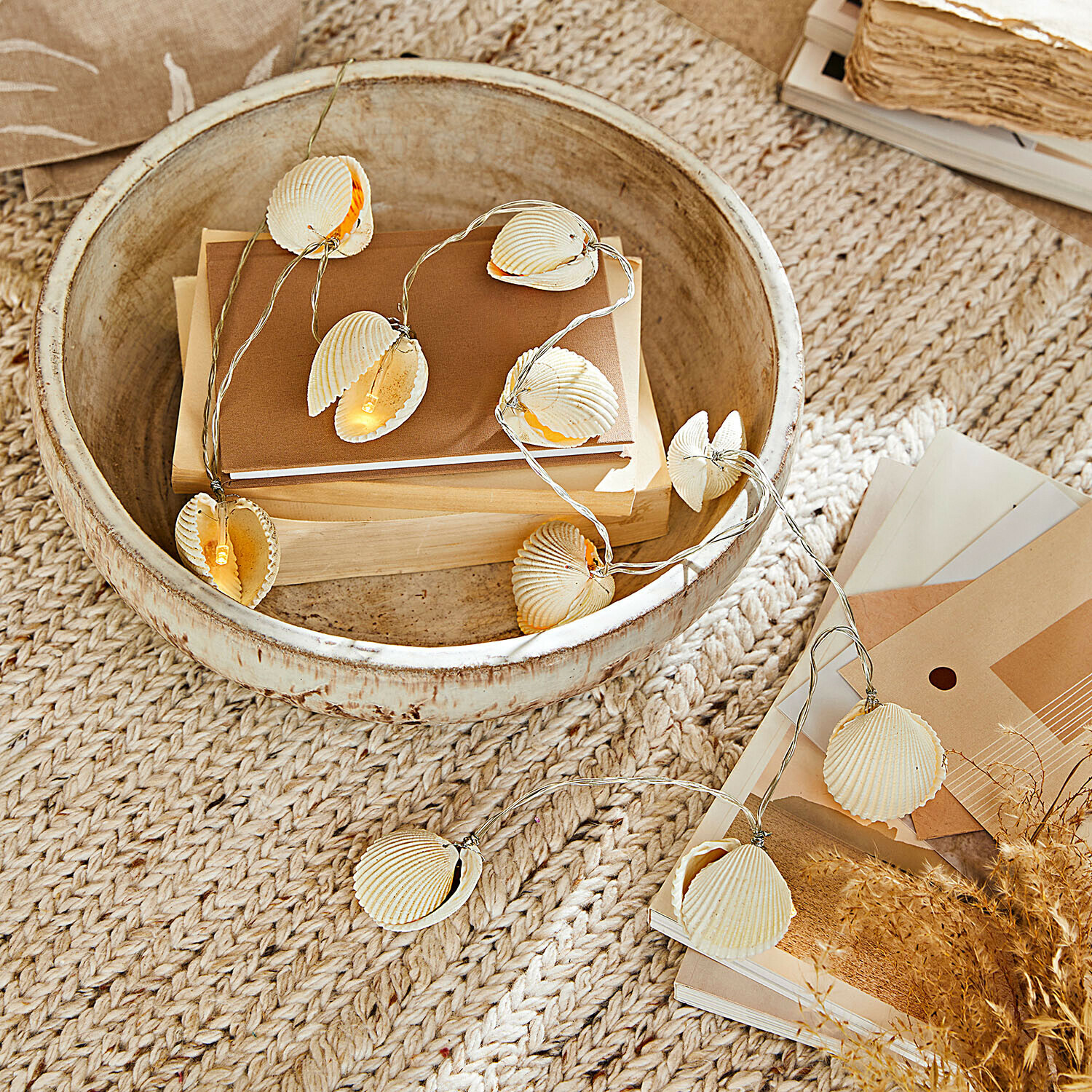 Světelný řetěz s přírodními mušlemi na béžovém pozadí s knihami a dekoracemi v rustikální misce