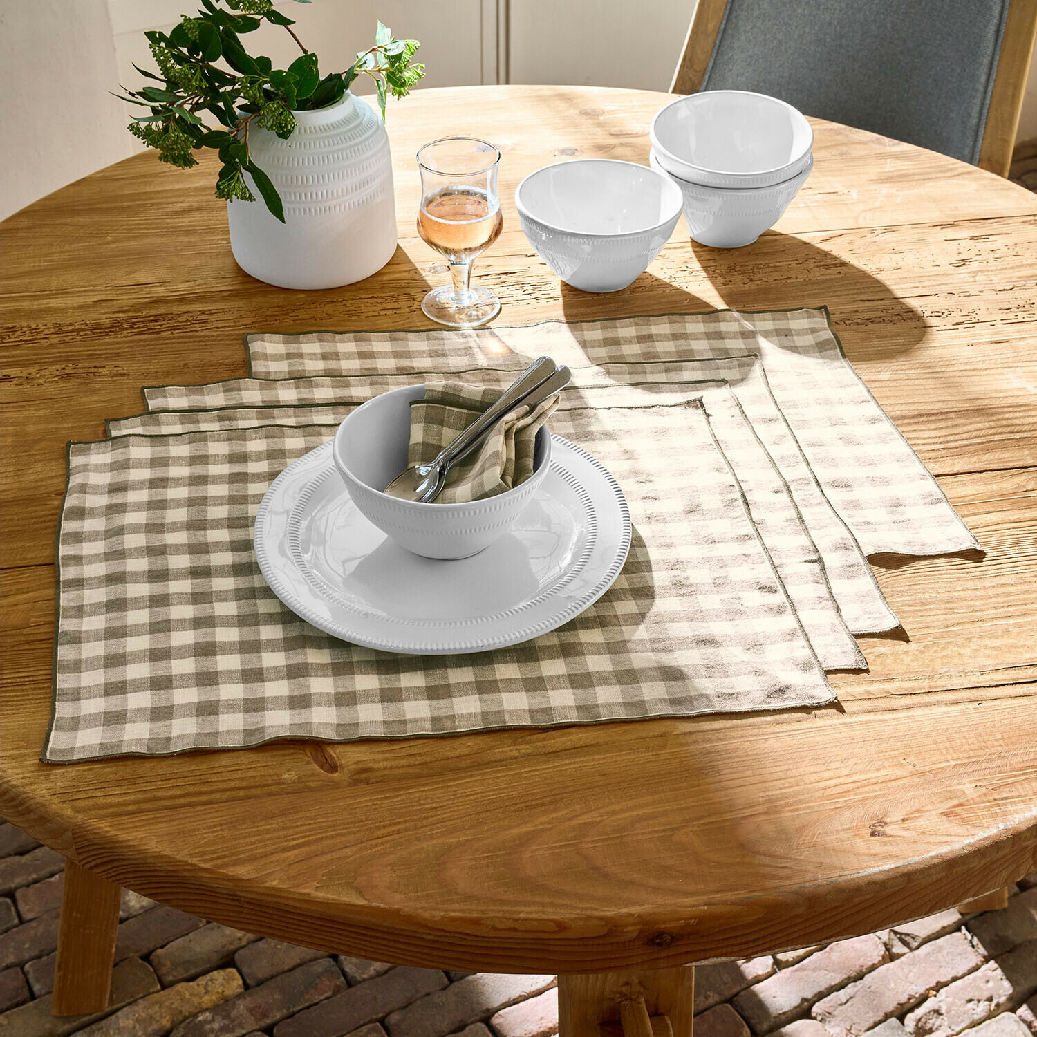 Bílé keramické misky a talíř na dřevěném stole s béžovo-bílými lněnými prostíráními v kostce