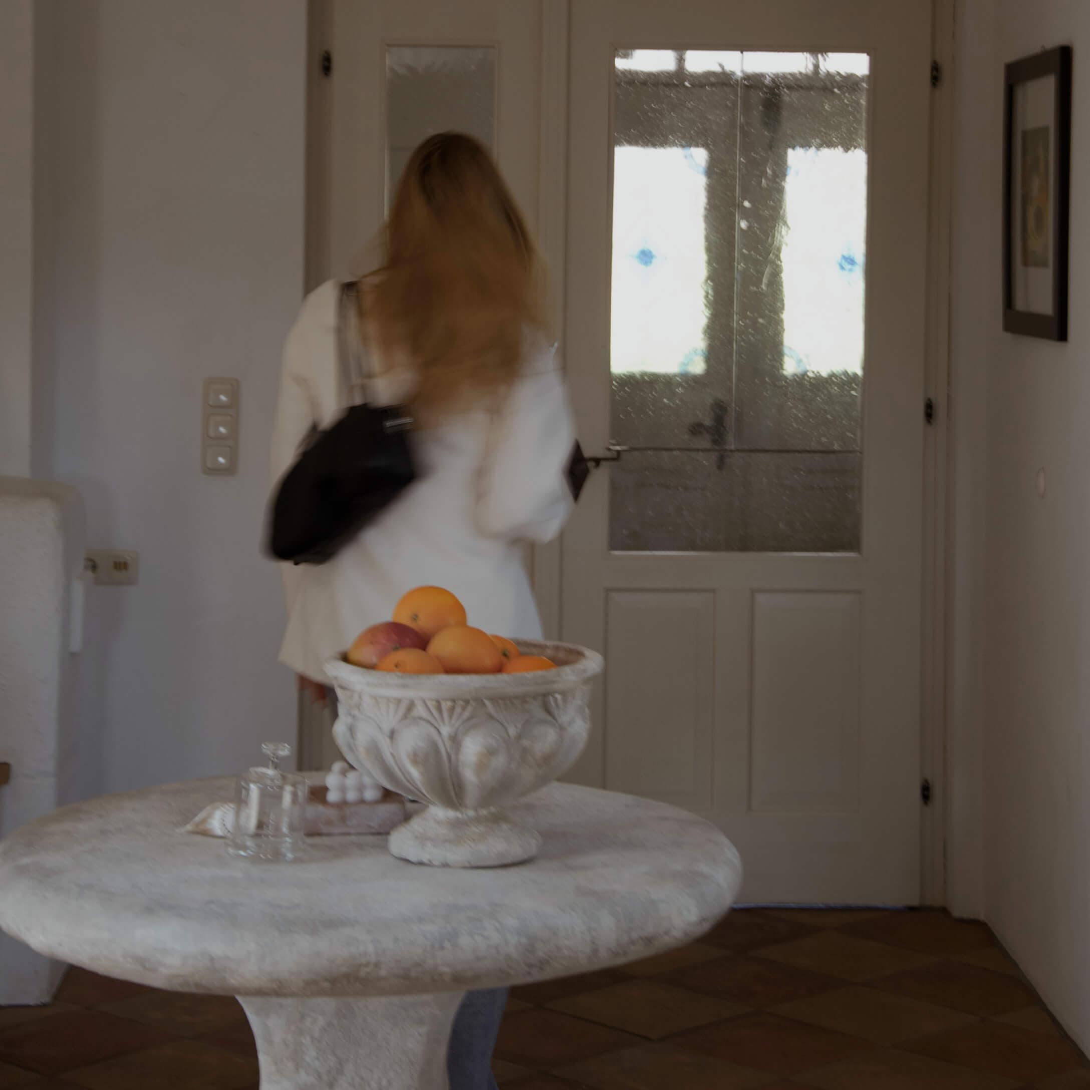 Dekorativní kamenná mísa s pomeranči na kulatém stole v světlém interiéru s prosklenými dveřmi.