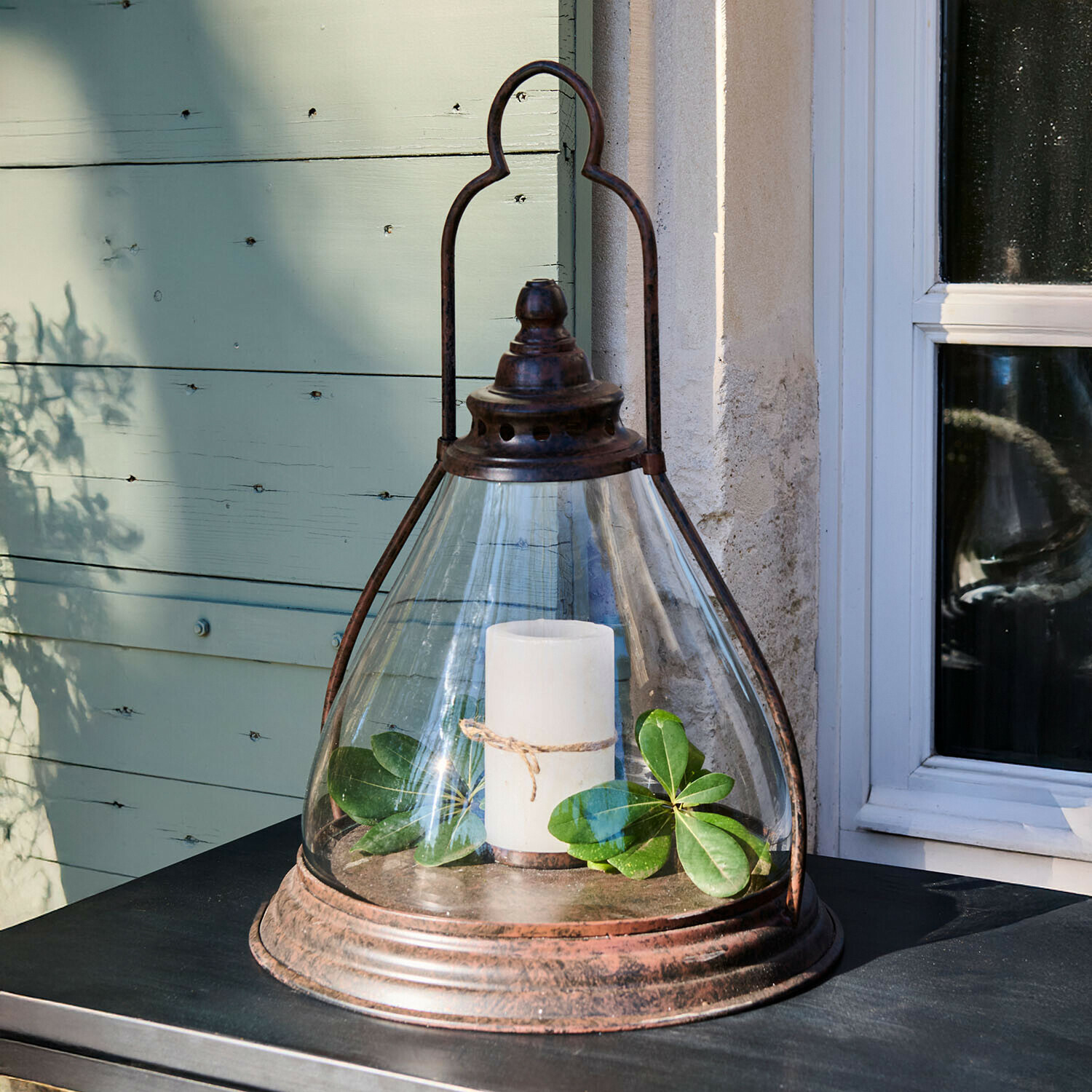 Skleněná lucerna s rezavým kovovým rámem, bílá svíčka a zelené listy na stole