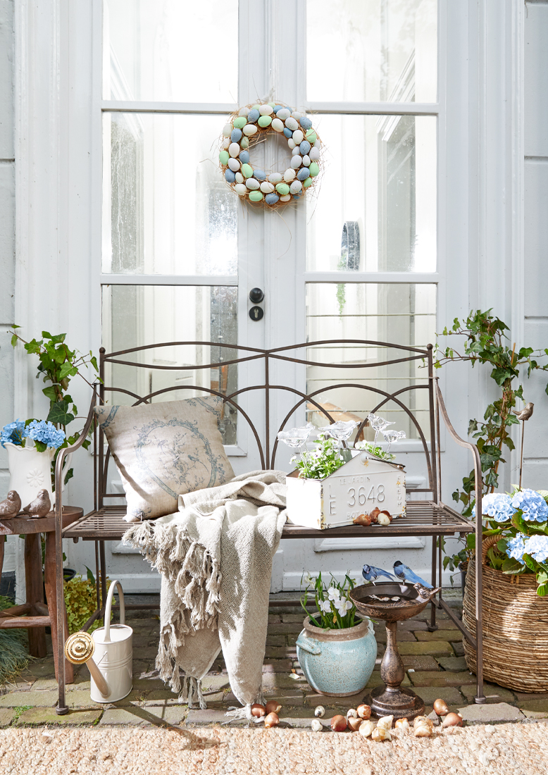 Kovová zahradní lavička s béžovou dekou a polštářem, květinové dekorace a věnec z pastelových vajec