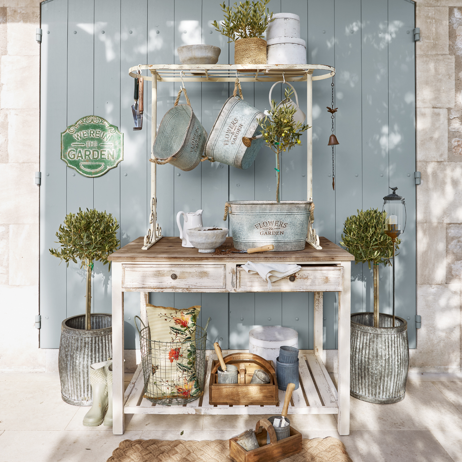 Bílý dřevěný zahradní stůl s kovovými květináči a zahradními nástroji před modrými dveřmi
