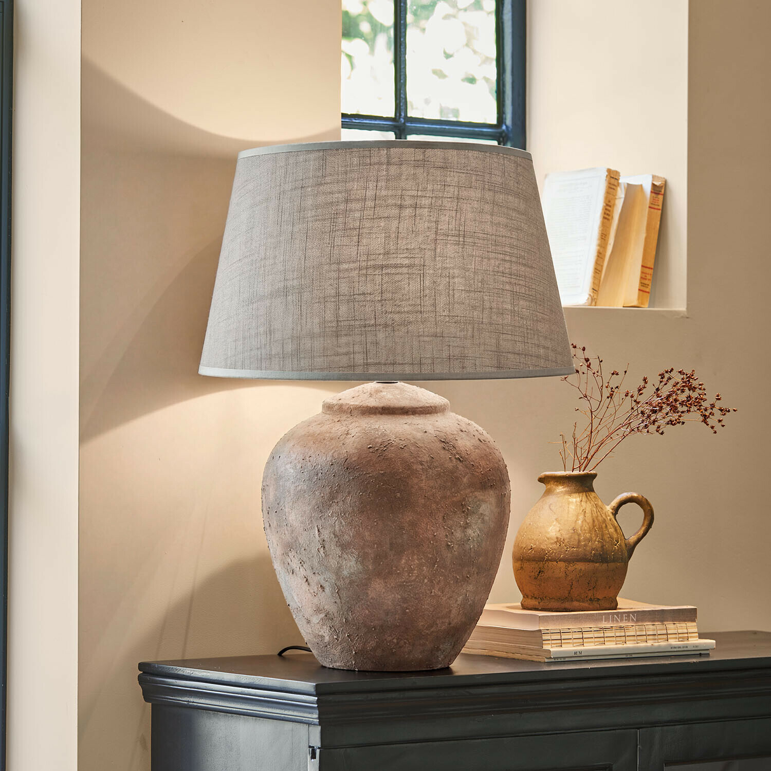 Stolní lampa Kelvador Stolní lampa s hliněnou základnou a šedým stínidlem na tmavém komodě v útulném interiéru