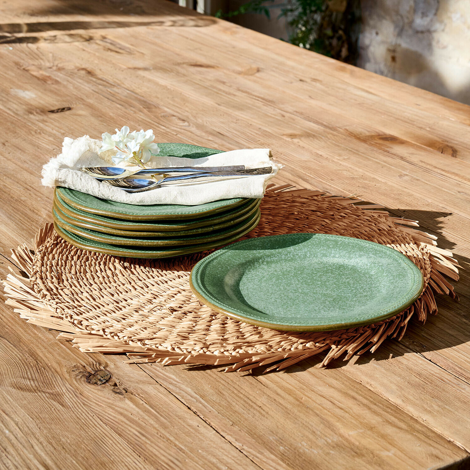 Sada zelených keramických talířů na pleteném prostírání na dřevěném stole s kovovým příborem