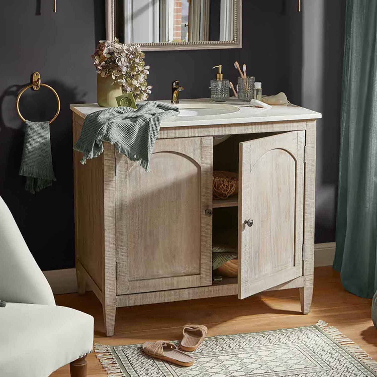 Světle dřevěná koupelnová skříňka s bílým umyvadlem a zlatou baterií, zelené ručníky a dekorace