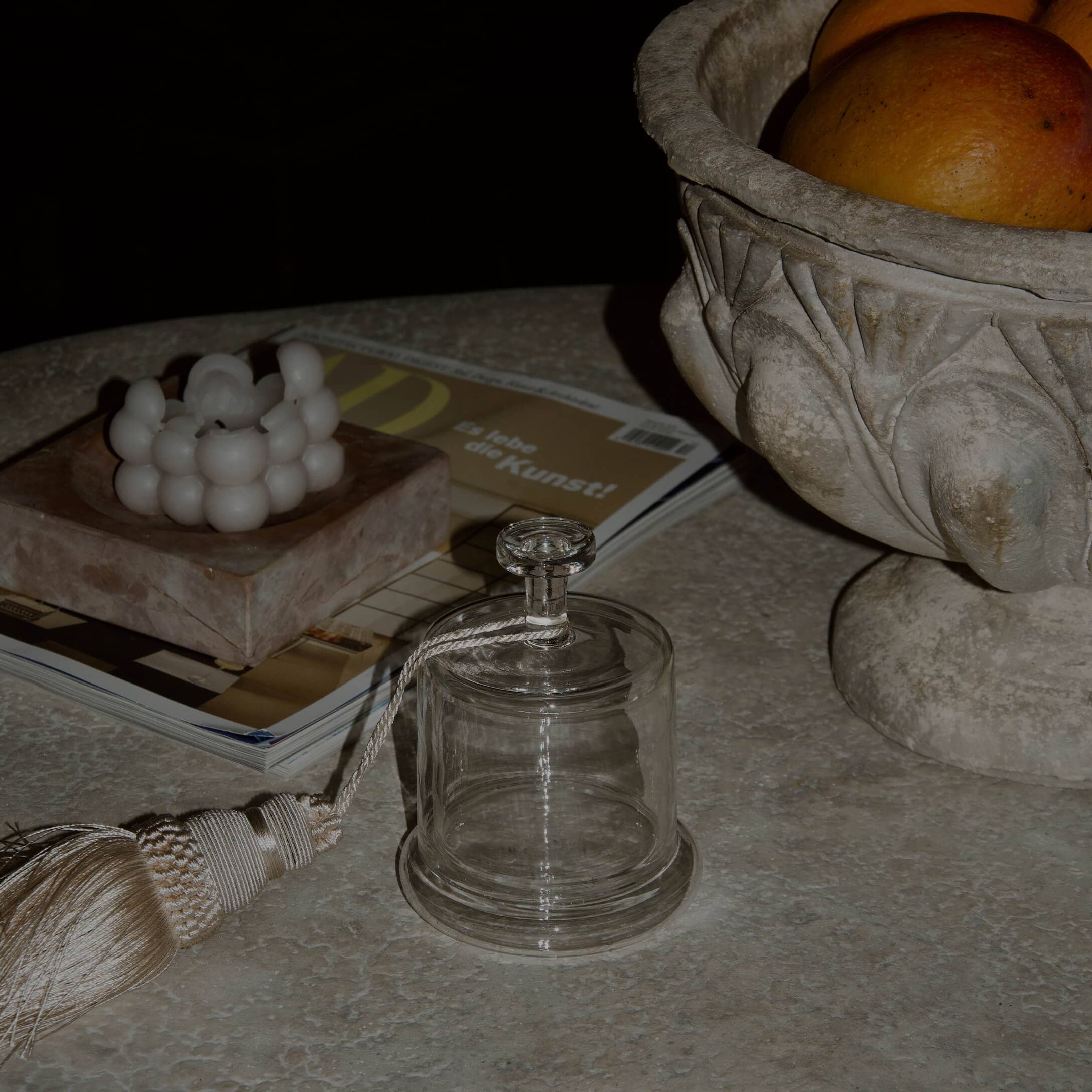 Průhledný skleněný poklop s ozdobným střapcem na mramorovém stole s dekorativní svíčkou a kameninovou miskou