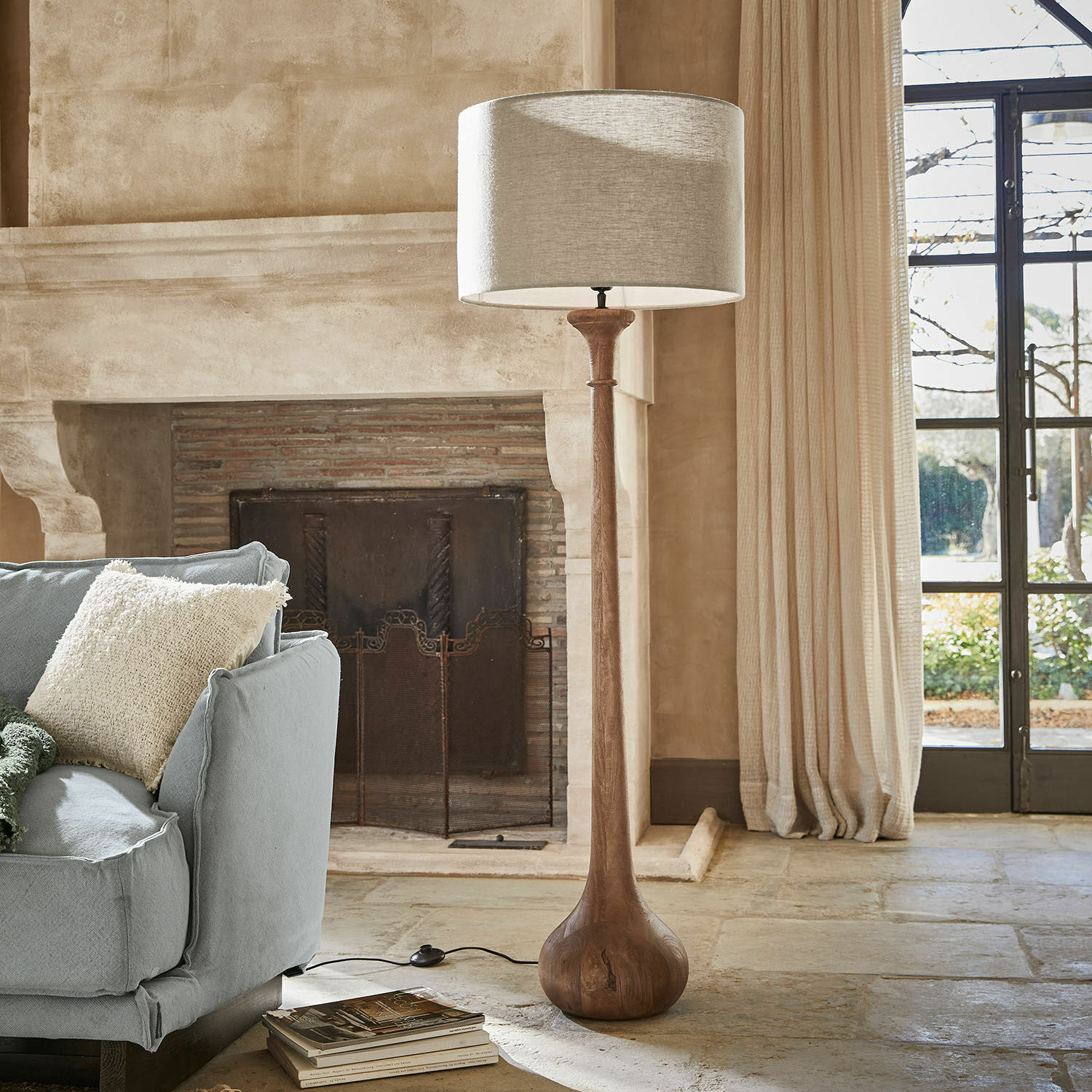 Stojací lampa Andrées Stojací lampa s dřevěnou základnou a béžovým stínidlem v útulném obývacím pokoji s krbem