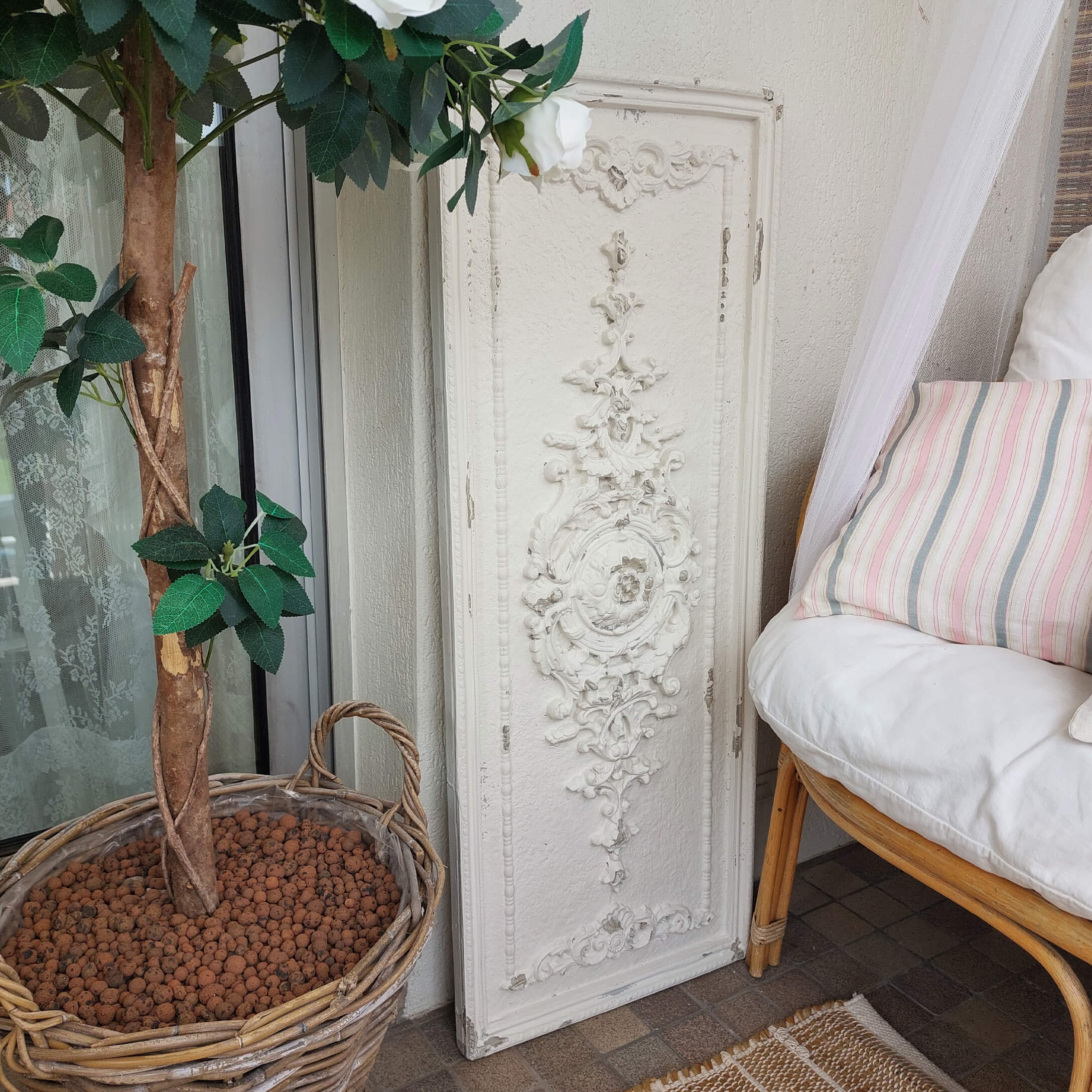 Bílý dekorativní nástěnný panel s vintage ornamentem vedle proutěného koše s rostlinou