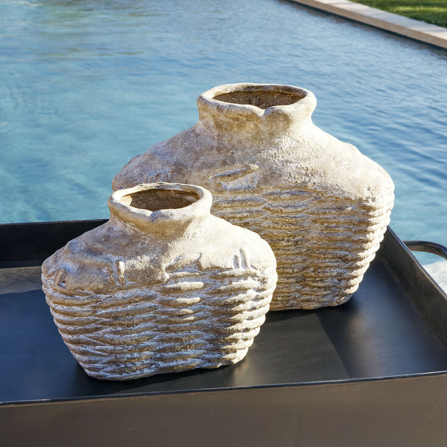 Dvě dekorativní kamenné vázy v béžové barvě na černém podnosu u bazénu