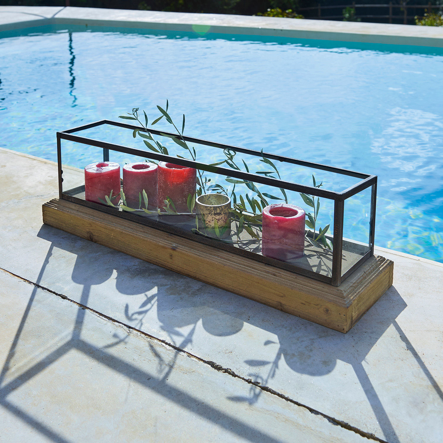 Skleněná dekorace s kovovým rámem a dřevěným podstavcem, červené svíčky a zelené větvičky uvnitř