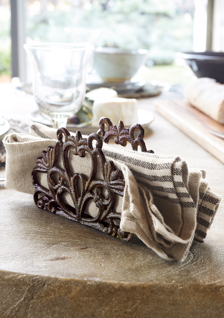 Rustikální kovový stojan na ubrousky s ozdobným vzorem v rezavé barvě a lněný ubrousek na kamenném stole