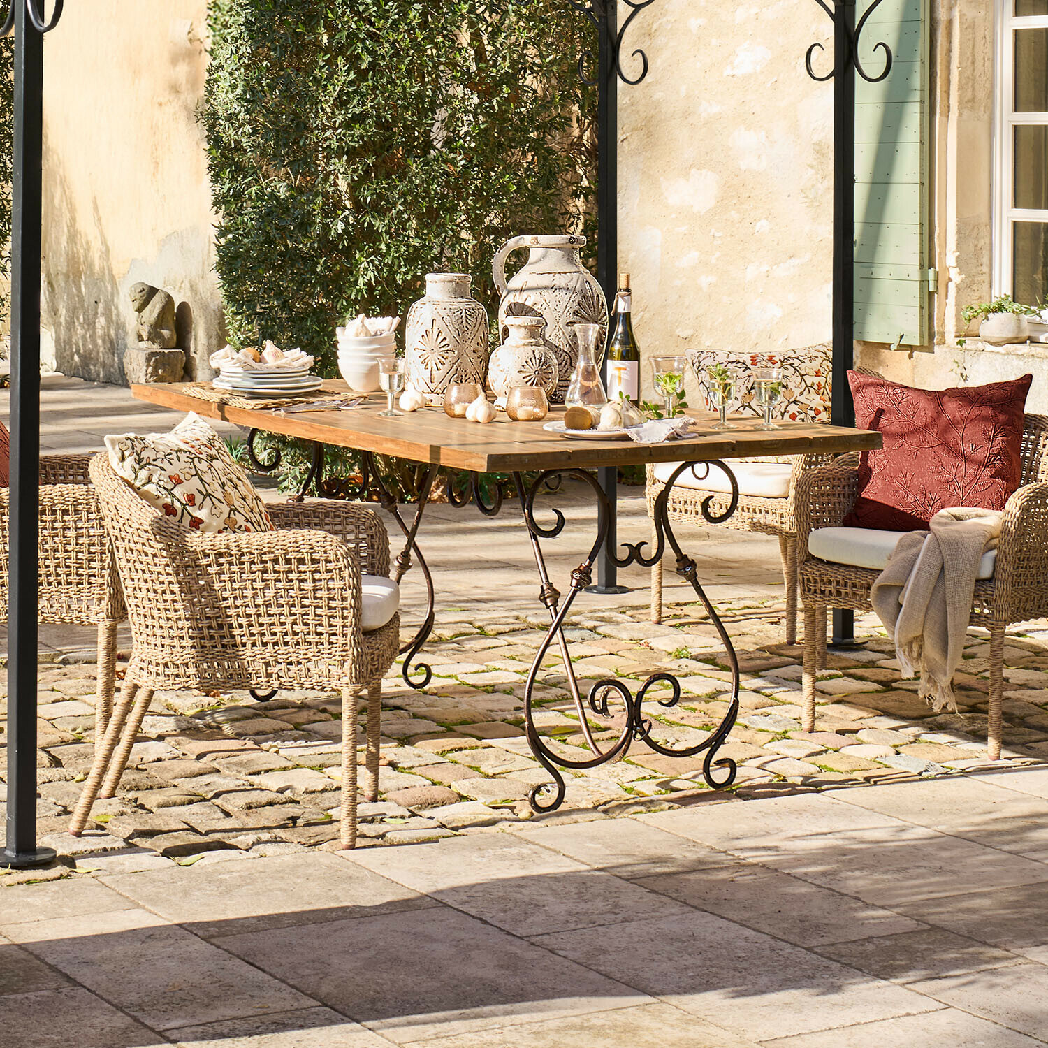 Zahradní stůl s dřevěnou deskou a kovovými nohami, ratanové židle s polštáři a keramické dekorace