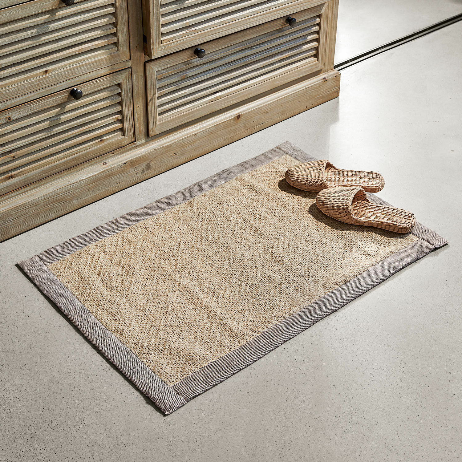 Předložka do koupelny Wilder Béžový koberec z přírodního materiálu s šedým lemem na světlé podlaze vedle dřevěné skříňky
