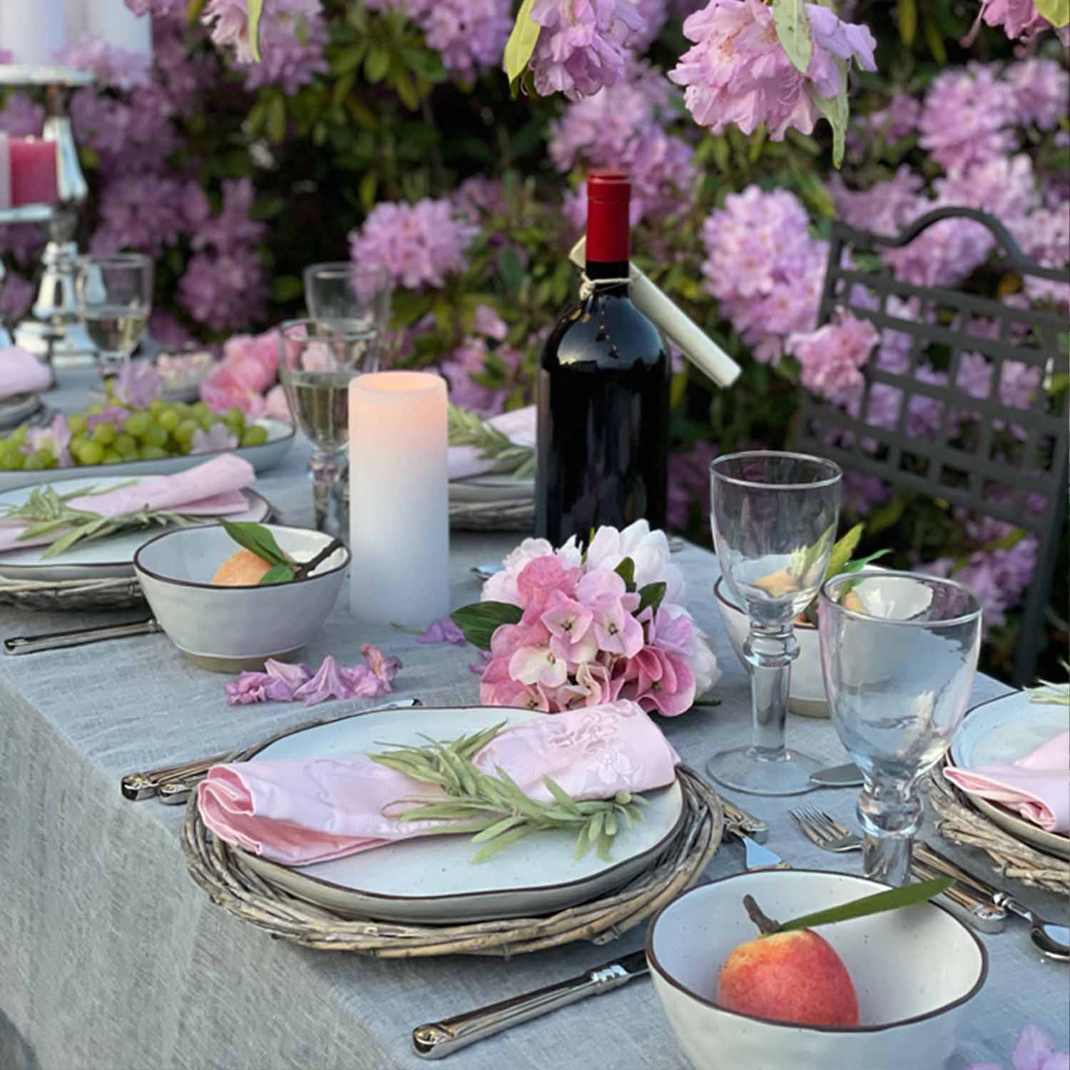 Elegantně prostřený stůl s bílým nádobím, růžovými ubrousky a červeným vínem v květinové zahradě