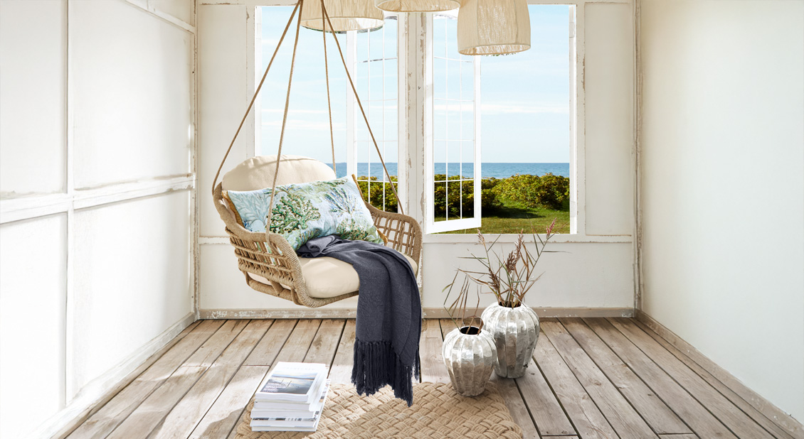 Závěsné proutěné křeslo s světlým polstrováním a dekorativním polštářem v námořním stylu v světlé místnosti s výhledem na moře