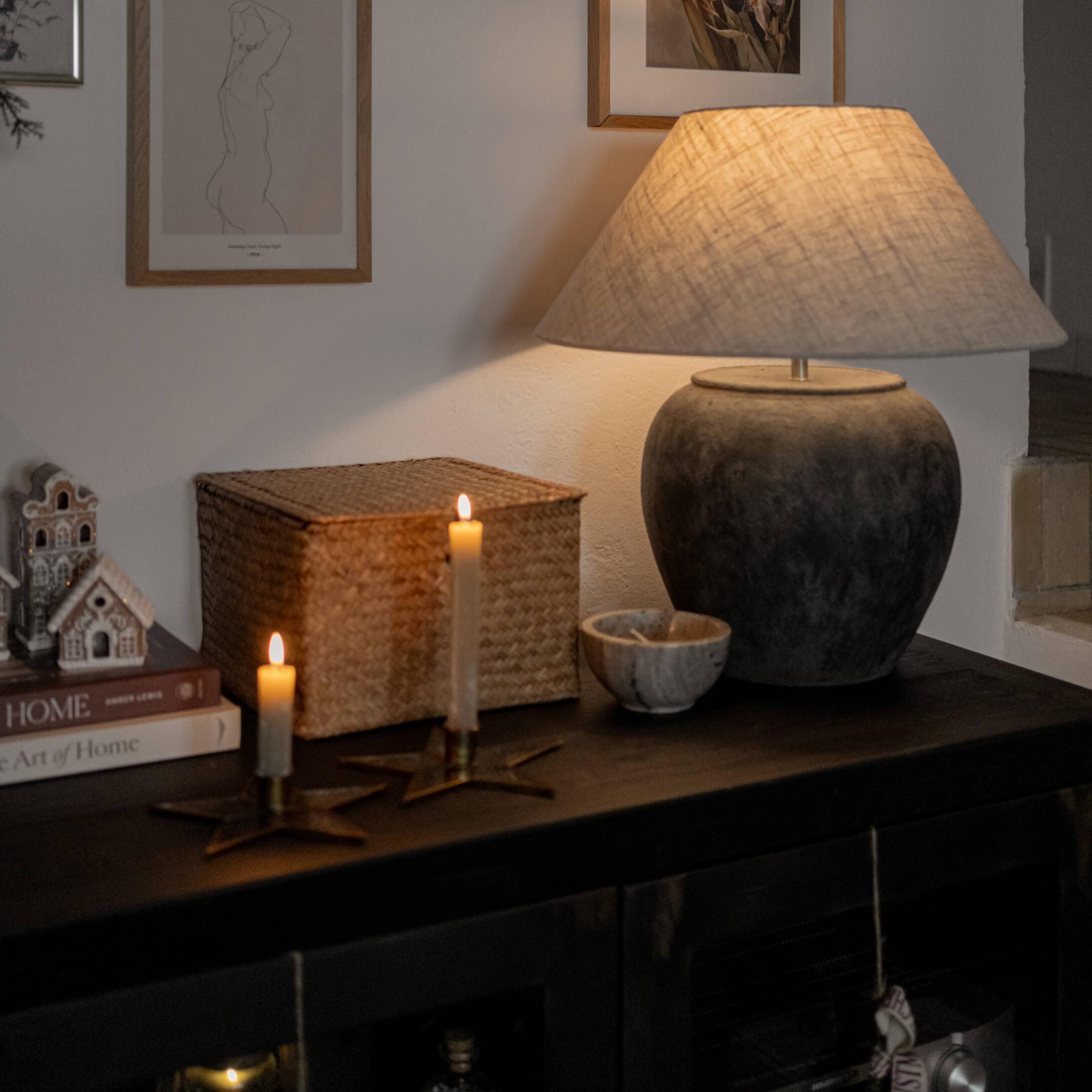 Stolní lampa z tmavě šedé keramiky s béžovým stínidlem na tmavém nábytku se svíčkami a dekoracemi
