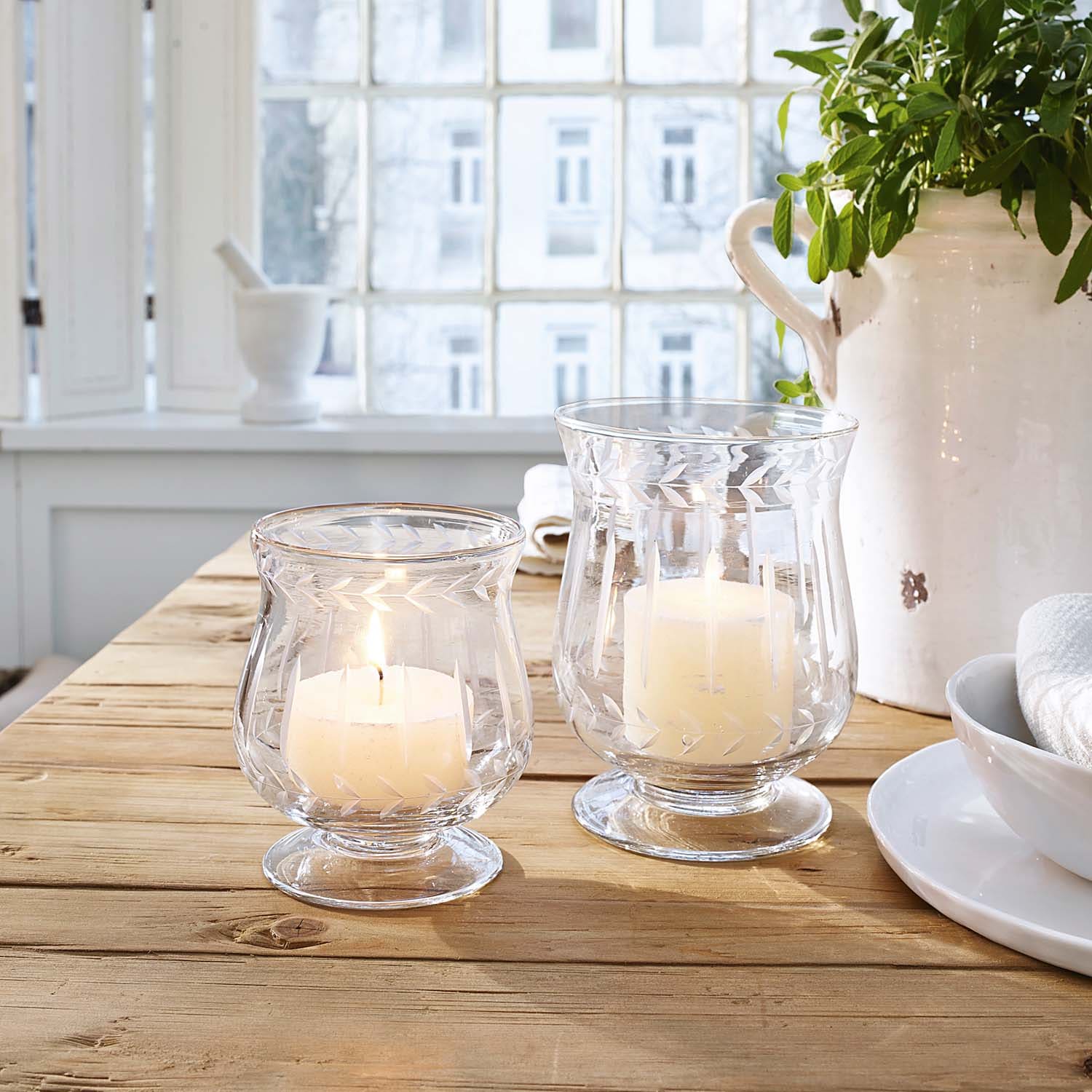 Dva skleněné svícny s bílými svíčkami na dřevěném stole v světlé místnosti