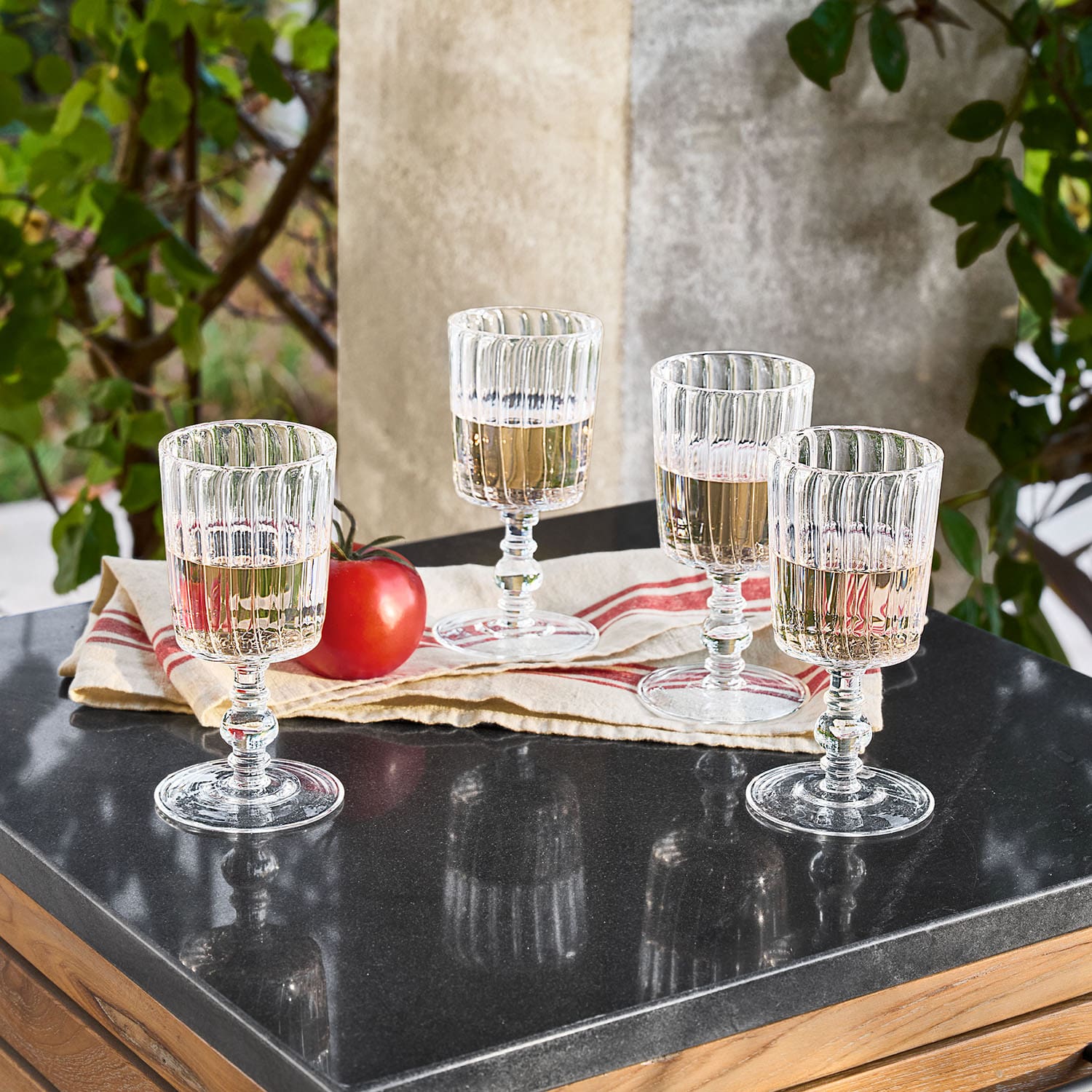 Čtyři žebrované sklenice z čirého skla s bílým vínem na černém stole vedle rajčete