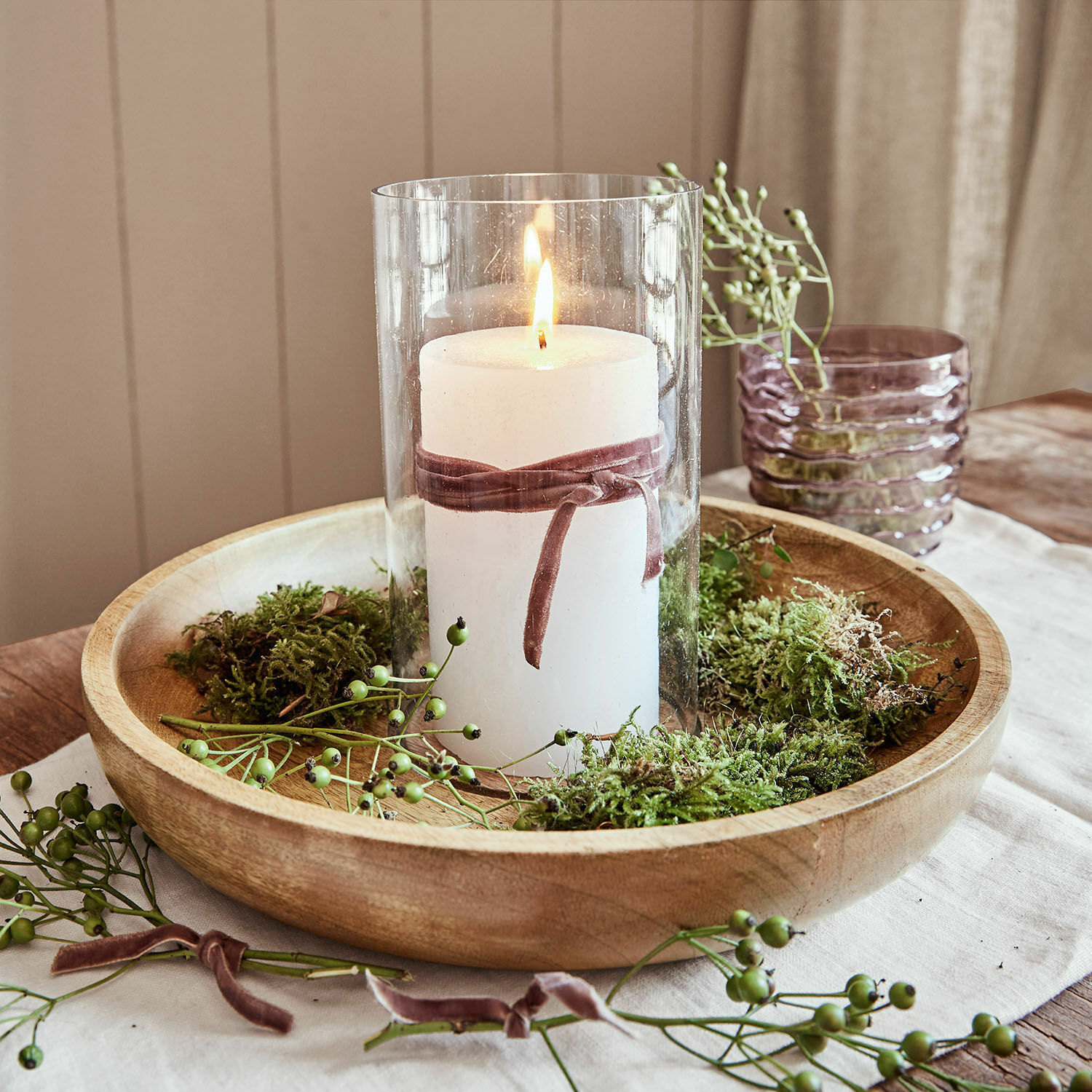 Bílá svíčka ve skleněném válci s sametovou stuhou na dřevěném podnosu s mechem