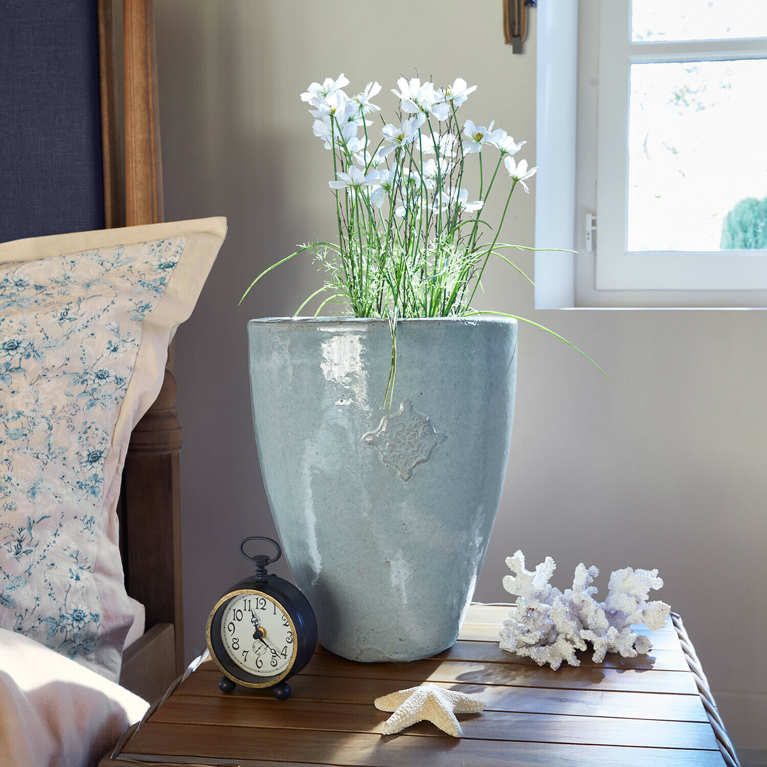 Šedá keramická květináč s bílými květy na dřevěném stolku vedle budíku a mořských dekorací