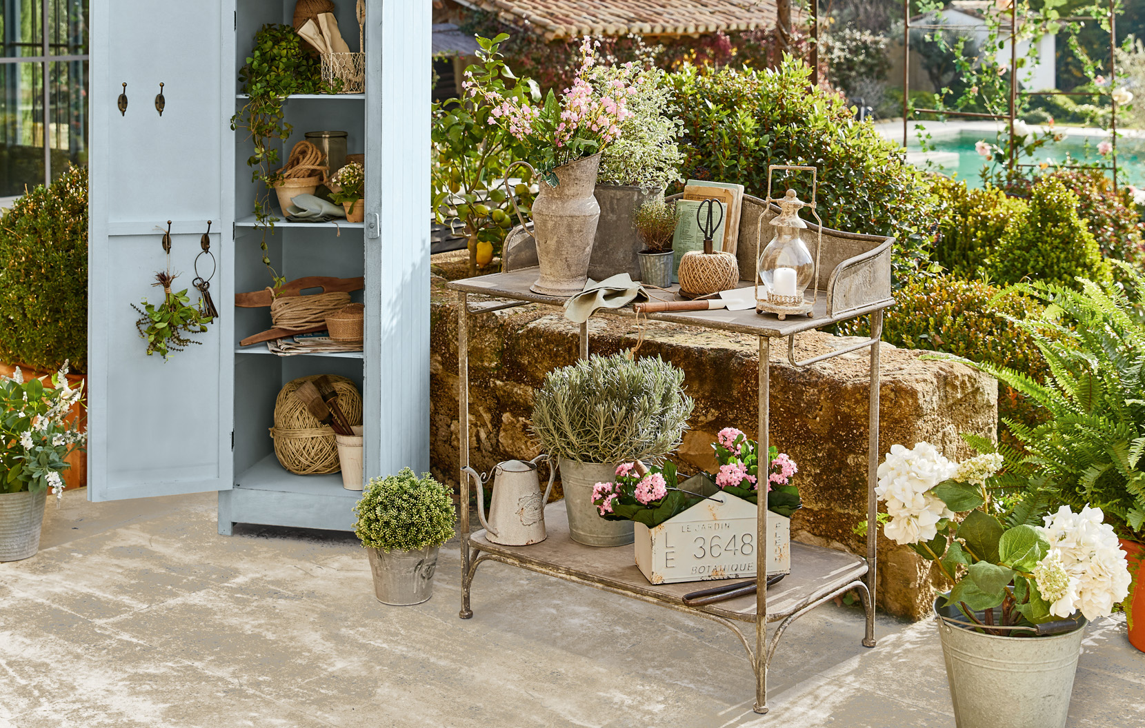 Kovový zahradní vozík se shabby chic dekoracemi a květinami na terase u modré skříně.
