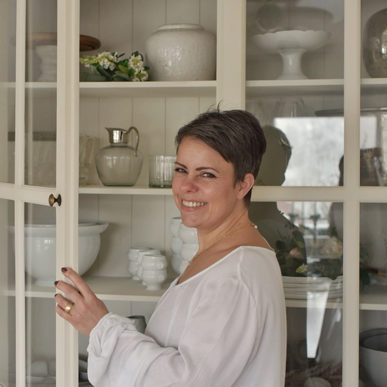 Žena v bílém topu otevírá krémovou skříňku s porcelánem a dekoracemi doma