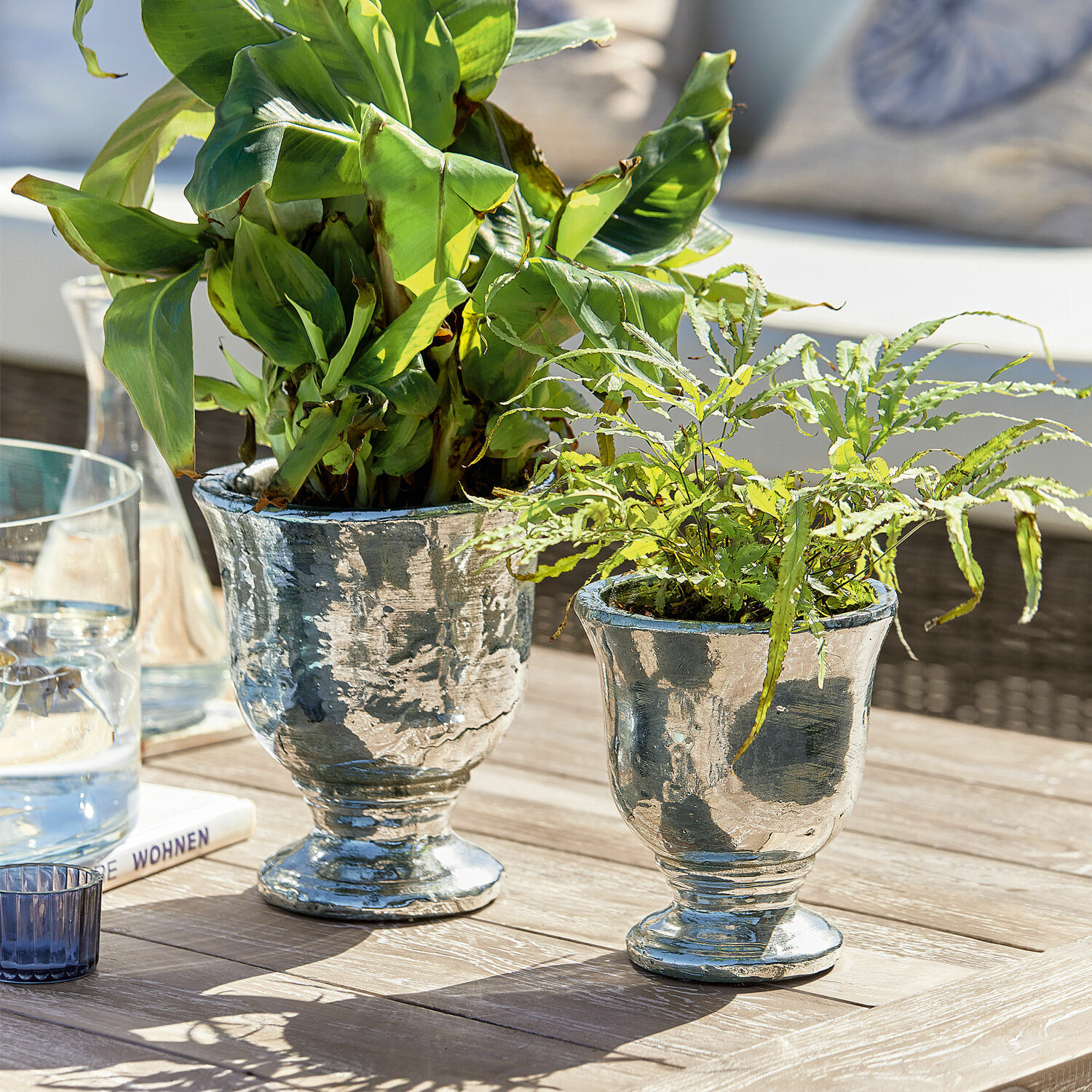 Dvě stříbrné kovové květináče se zelenými rostlinami na dřevěném stole v jasném prostředí