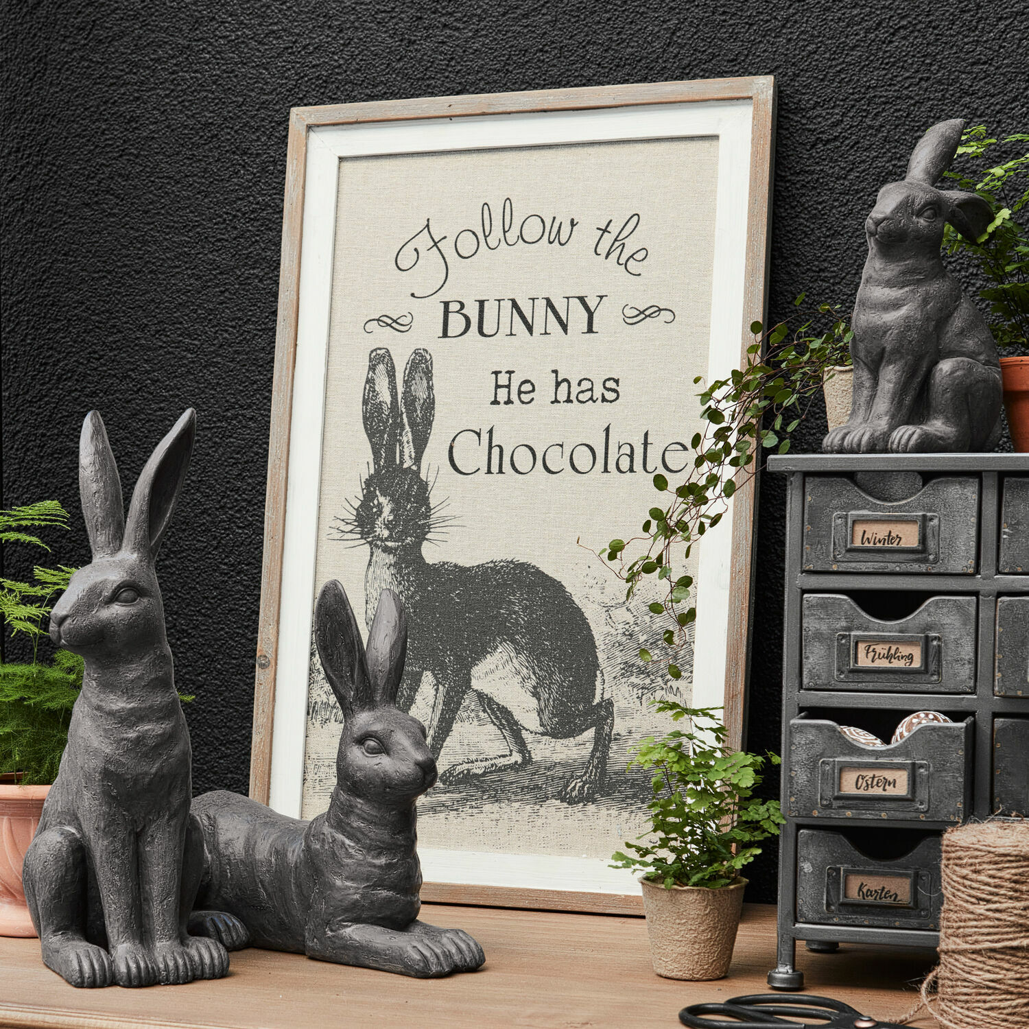 Obraz Chocolate Šedé dekorativní sošky zajíců s rámem s nápisem o zajíci a čokoládě na dřevěném stole