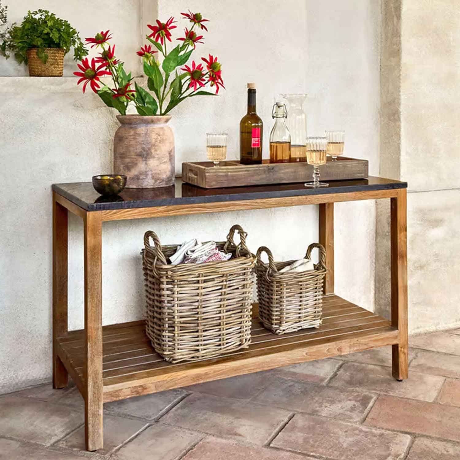 Dřevěný stolek s černou deskou, váza s červenými květy a proutěné koše pod ním v rustikálním interiéru