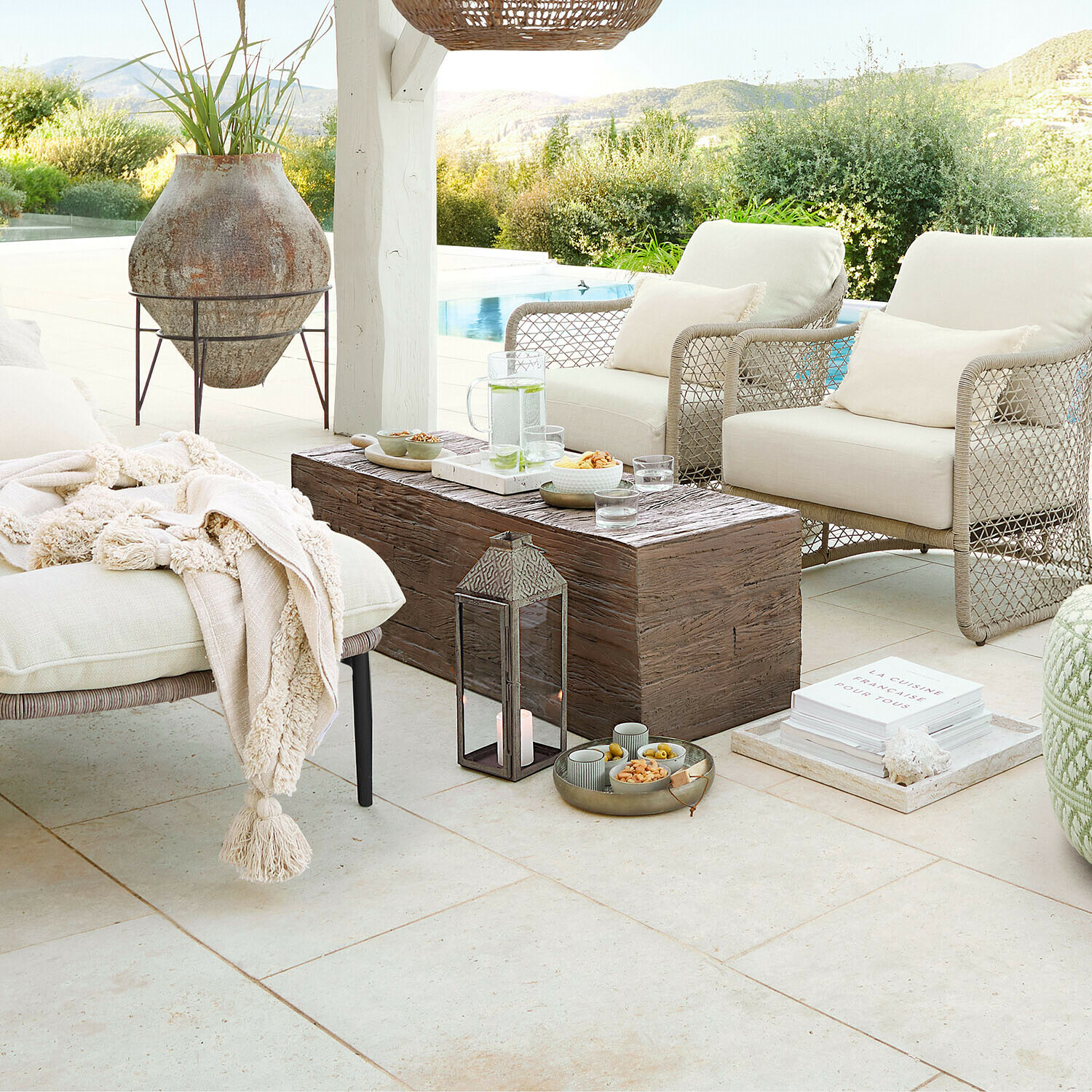 Pohodlný zahradní nábytek s béžovými polštáři a dřevěným stolem na terase s výhledem na bazén a přírodu