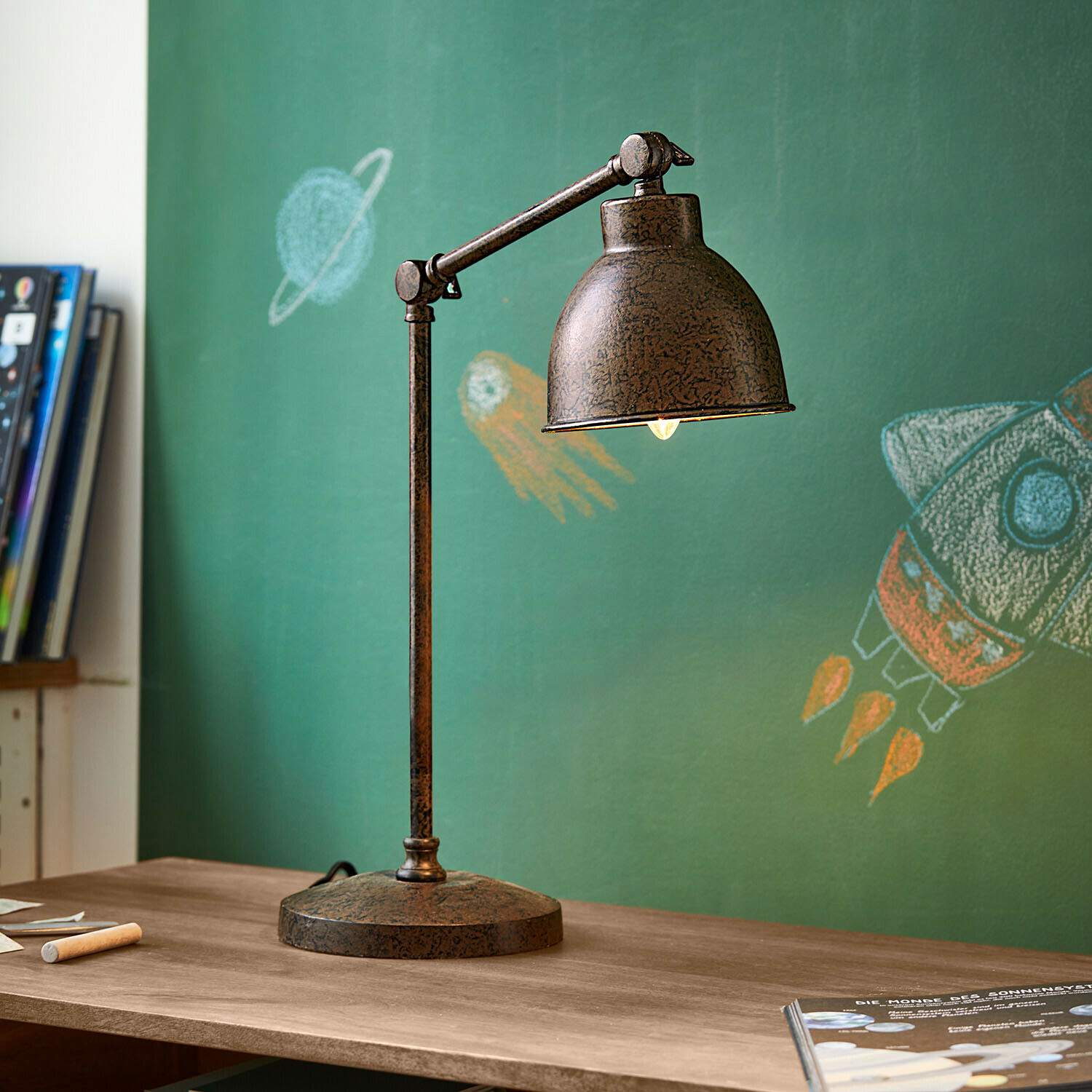 Stolní lampa Cheeklee Stolní lampa z kovu s rezavým efektem na dřevěném stole před zelenou tabulí s vesmírnými kresbami