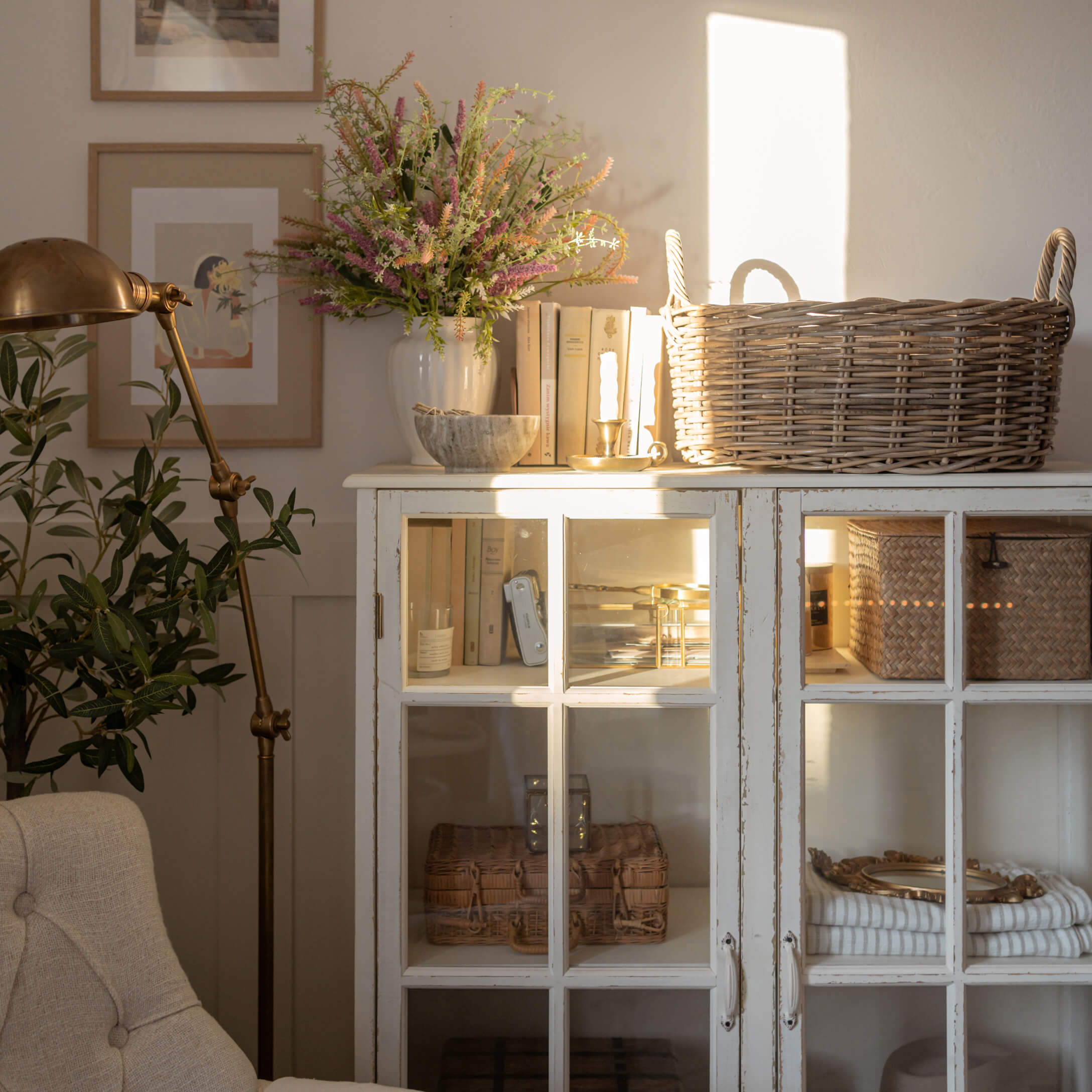 Bílá dřevěná skříňka se skleněnými dveřmi, proutěný koš a dekorace v světlém obývacím pokoji