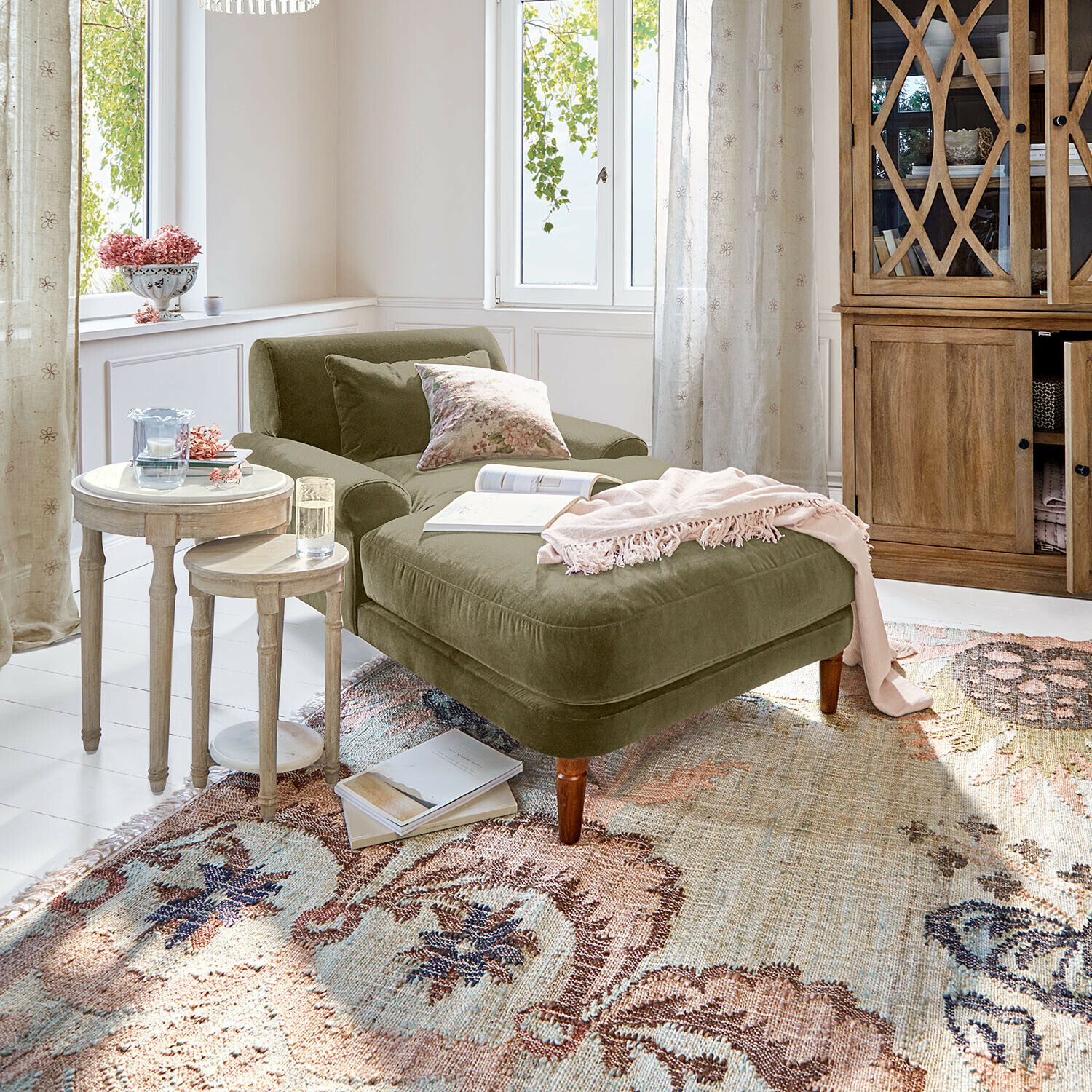 Zelené sametové lenoškové křeslo s polštáři a růžovou dekou v světlém obývacím pokoji s dřevěným nábytkem