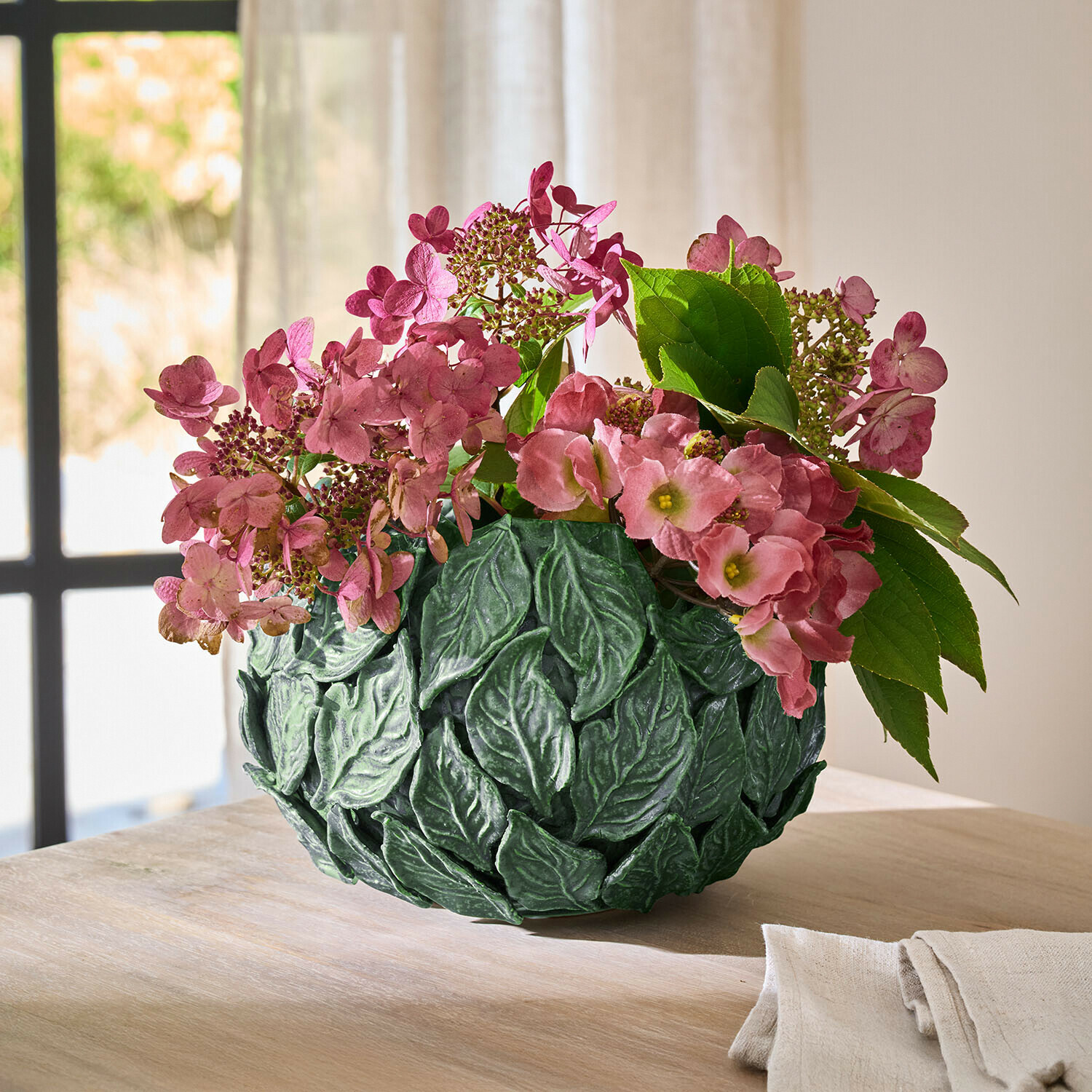 Zelená váza s listy a růžovými květy na dřevěném stole v světlé místnosti jako dekorace