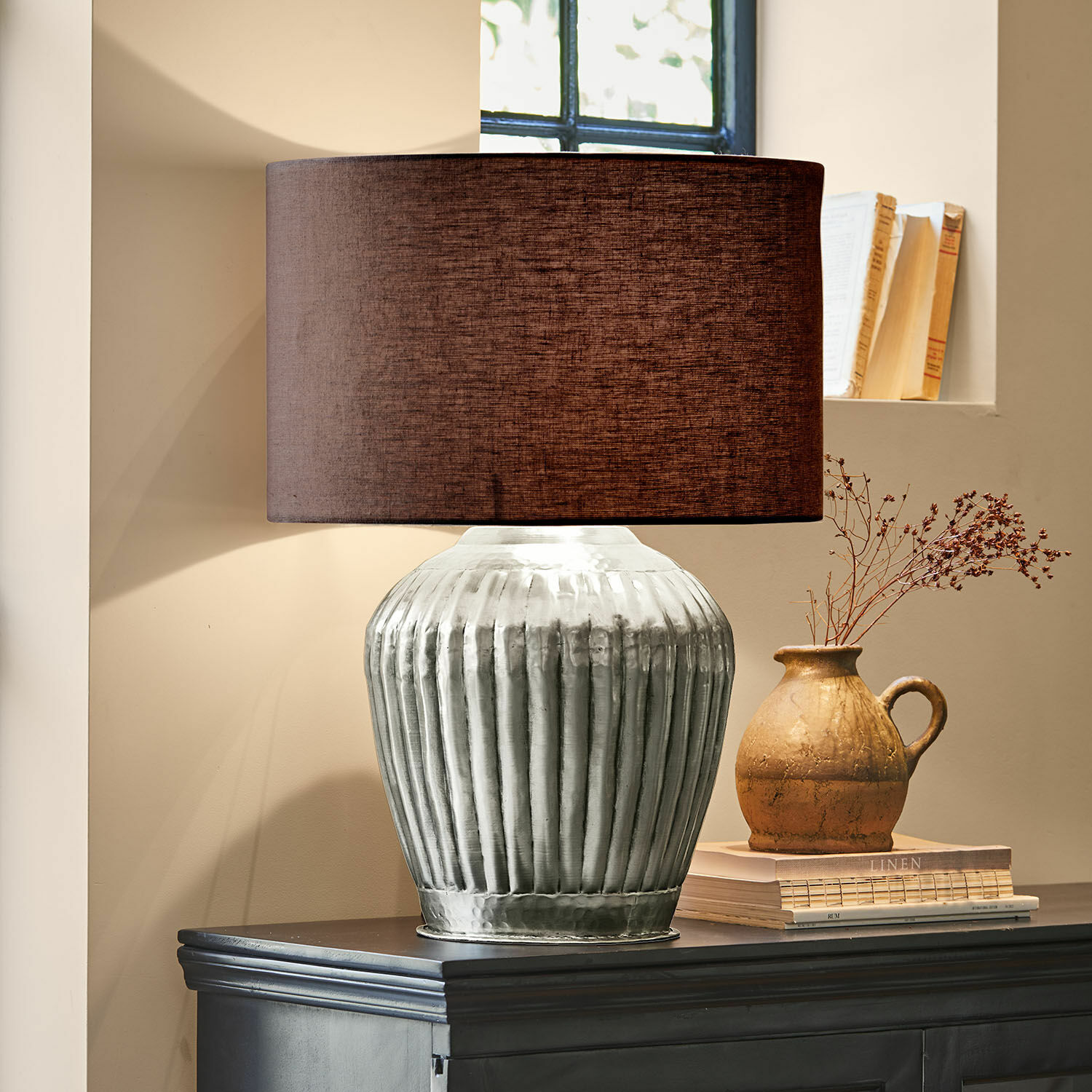 Stolní lampa Fauville Stolní lampa se stříbrnou kovovou základnou a hnědým stínidlem na tmavé komodě